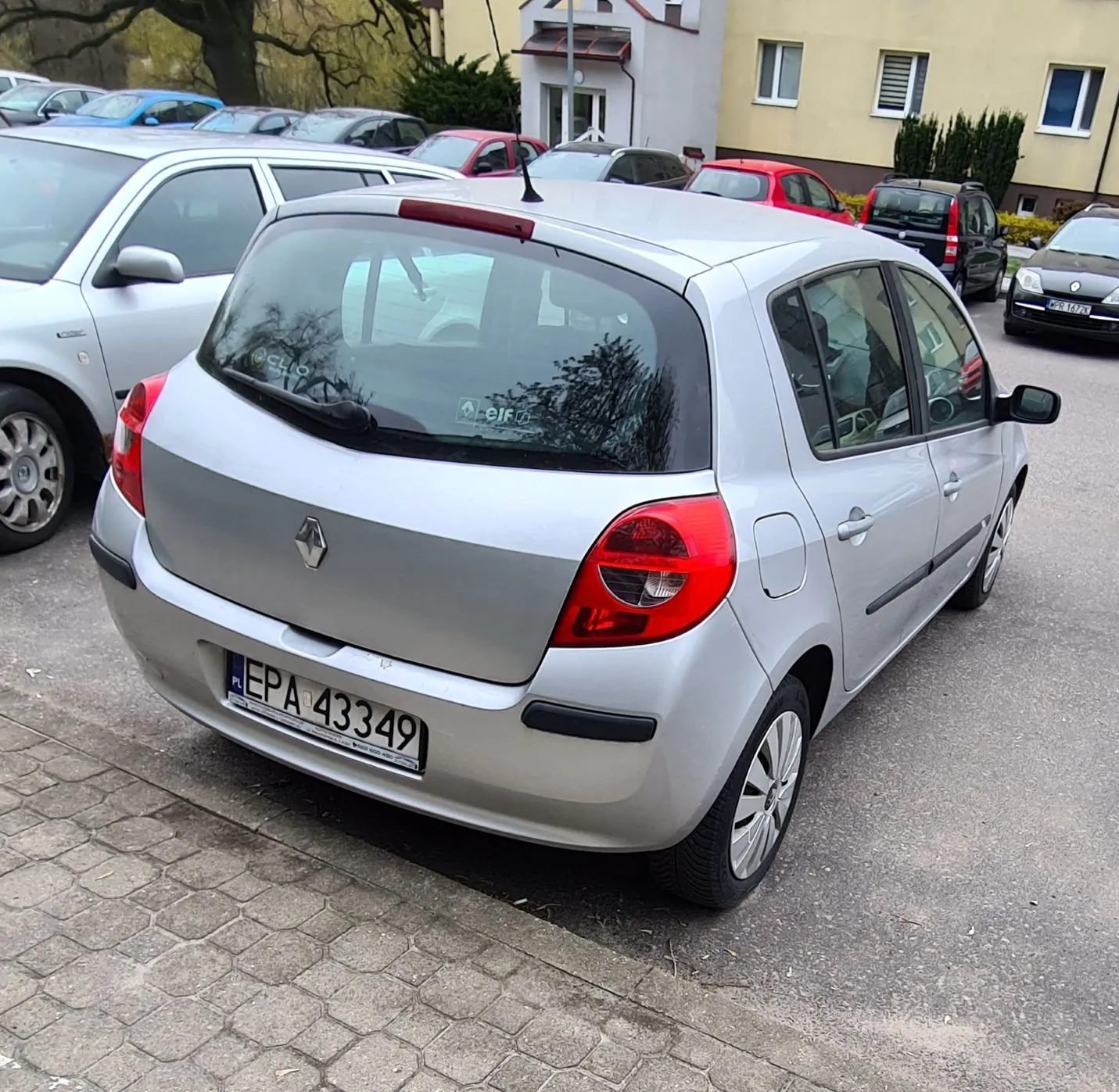 Srebrny samochód marki Renault z tablicą rejestracyjną EPA 43349 (PL) zaparkowany na placu parkingowym. W tle widoczne są inne pojazdy, budynki mieszkalne, drzewa oraz brukowana powierzchnia. Na szybie tylnej naklejki z napisami 'eif' i częściowo widocznym 'CLO'.