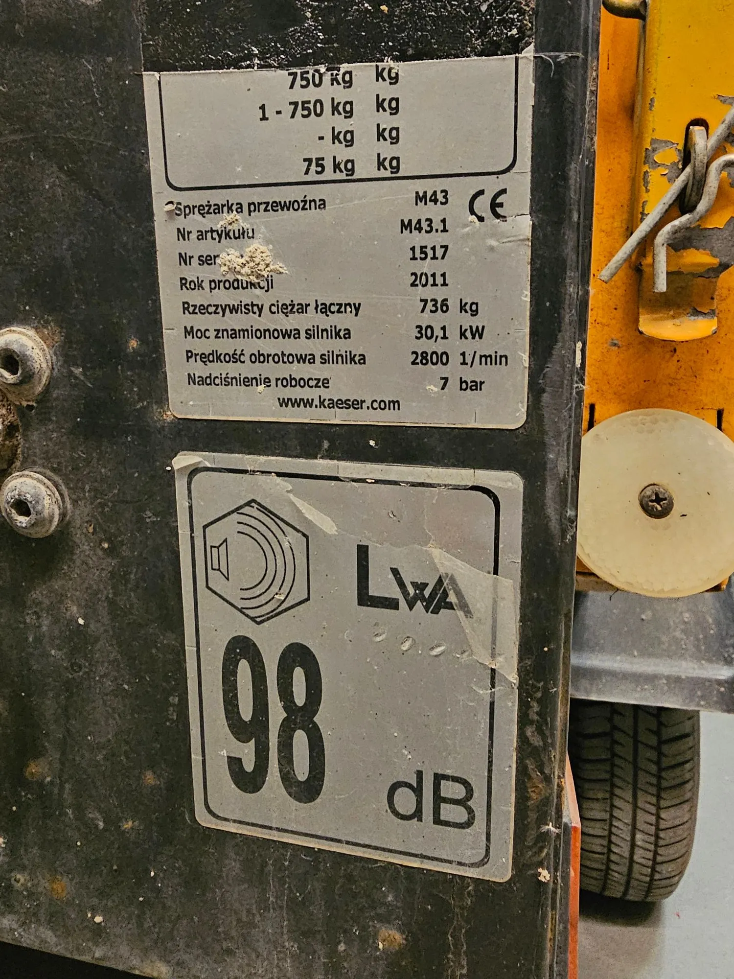 Zbliżenie na tabliczkę znamionową urządzenia przemysłowego. Tabliczka zawiera dane techniczne: 'Sprężarka przewozna M43 CE M43.1', 'Nr artykułu 1517', 'Nr ser. 2011', 'Rok produkcji 736 kg', 'Rzeczywisty ciężar łączny 30,1 kW', 'Moc znamionowa silnika 2800 1/min', 'Prędkość obrotowa silnika 7 bar', 'Nadciśnienie robocze'. Widnieje również adres strony www.kaeser.com. Po prawej stronie znajduje się osobna etykieta z oznaczeniem poziomu hałasu: 'LWA 98 dB'. Obraz jest zorientowany poziomo, ale tekst na tabliczkach jest obrócony o 90 stopni w lewo.