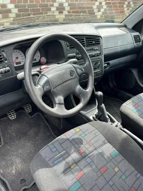 Wnętrze samochodu marki Volkswagen, widok z perspektywy pasażera. Widoczna jest czarna kierownica, deska rozdzielcza z zegarami, dźwignia zmiany biegów oraz dwa fotele z tkaninową tapicerką w szarym kolorze z kolorowymi, geometrycznymi wzorami w odcieniach czerwieni, żółci i niebieskiego. Na podłodze znajdują się czarne gumowe dywaniki.