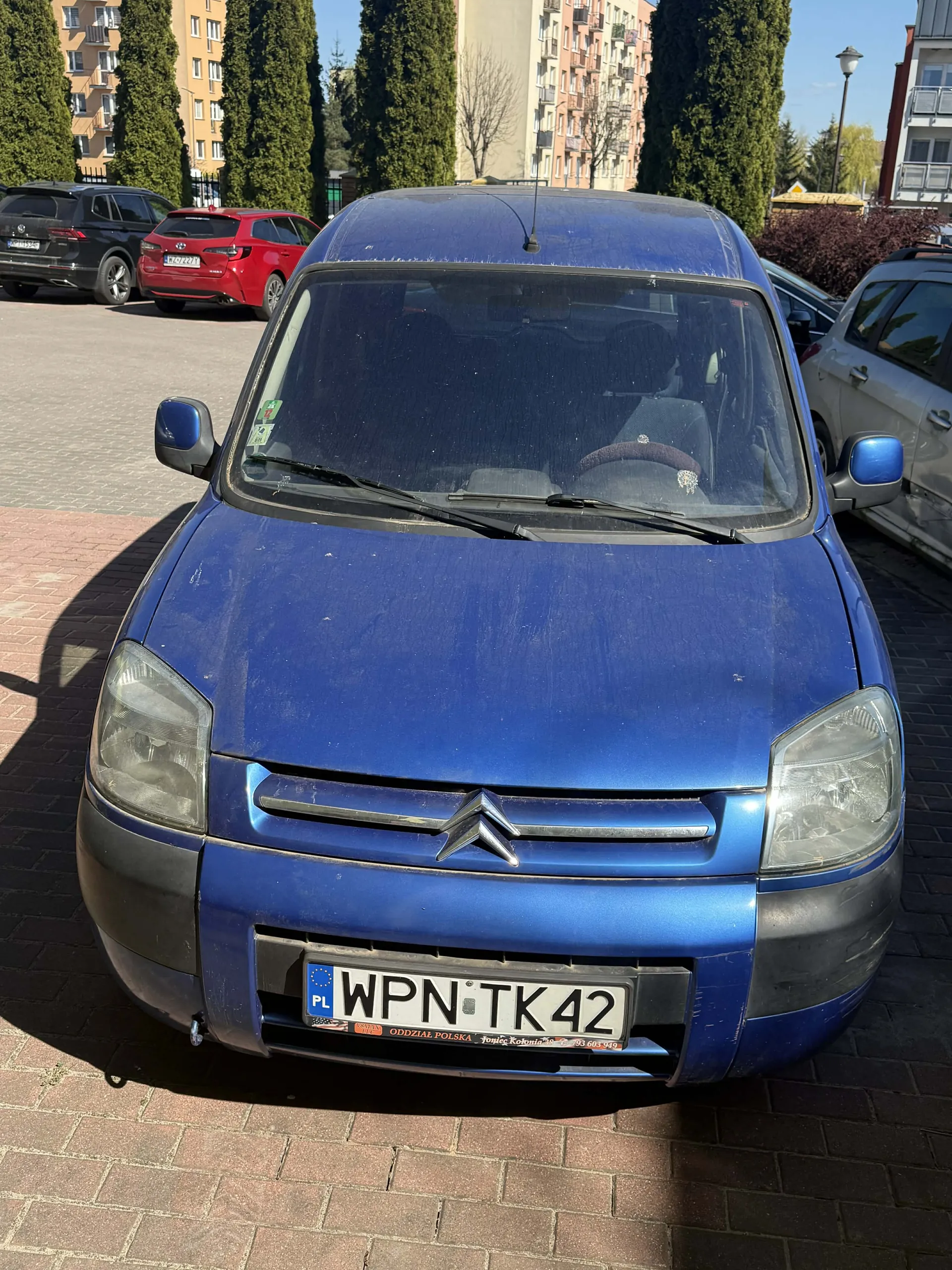 Przedni widok niebieskiego samochodu Citroën zaparkowanego na brukowanej powierzchni. Tablica rejestracyjna z numerem WPN TK 42 z kodem kraju PL. W tle widoczne inne pojazdy, w tym czerwony Toyota i czarny SUV. Scena obejmuje wielopiętrowe budynki mieszkalne, wysokie drzewa oraz jasne niebo. Na przedniej masce widoczny logo Citroën, reflektory i lusterka boczne.