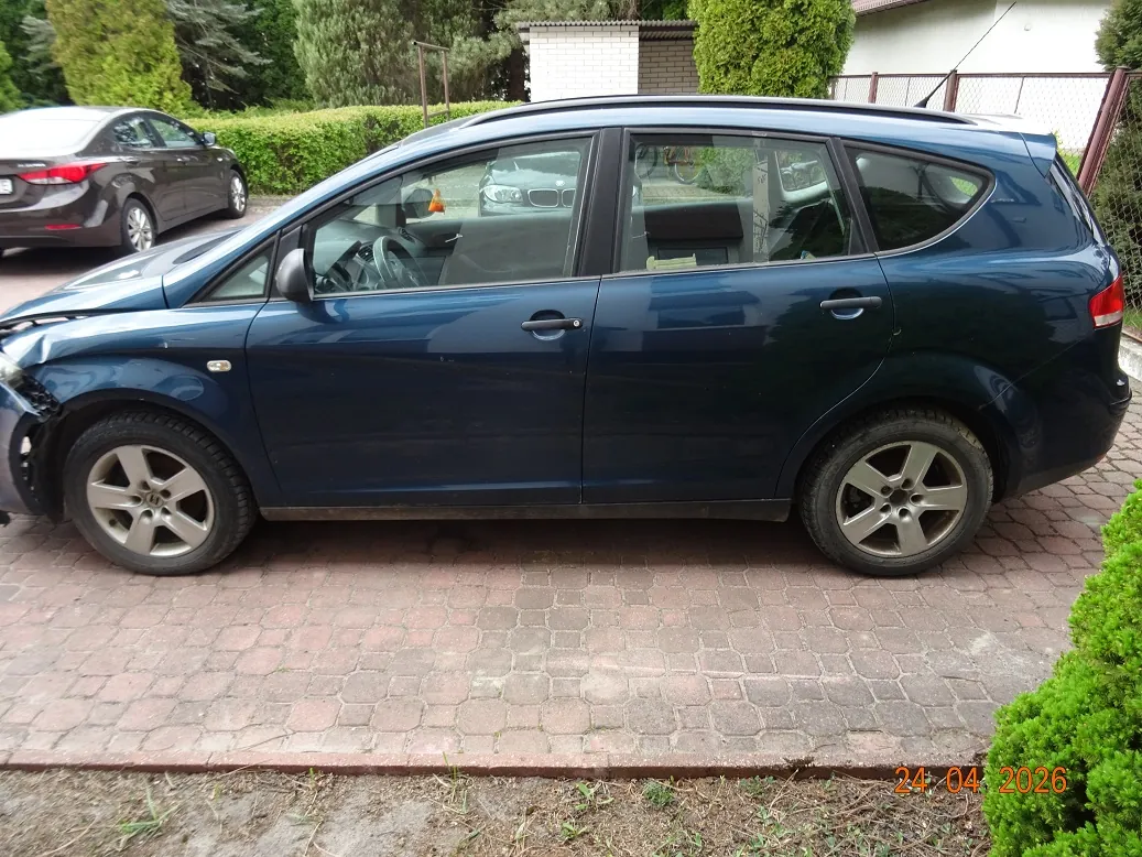 Profil ciemnoniebieskiego samochodu typu kombi zaparkowanego na kostce brukowej. Pojazd ma uszkodzony przedni zderzak po lewej stronie. W tle widoczny jest inny, brązowy samochód oraz zielone krzewy i drzewa. W prawym dolnym rogu znajduje się pomarańczowa data: 24.04.2026.
