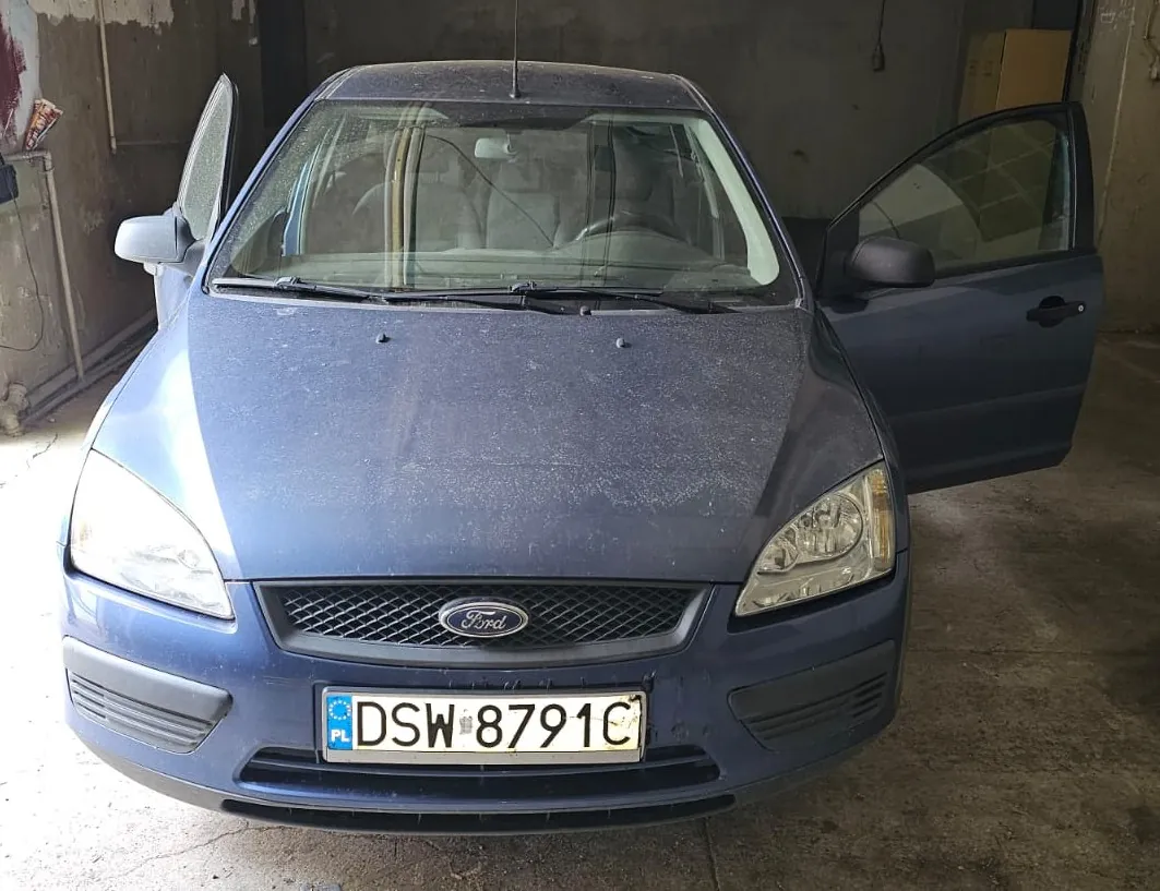 Niebieski samochód marki Ford zaparkowany w garażu. Pojazd ma otwarte przednie drzwi po obu stronach. Na masce widoczna jest warstwa kurzu. Samochód posiada polską tablicę rejestracyjną o numerze DSW 8791C.