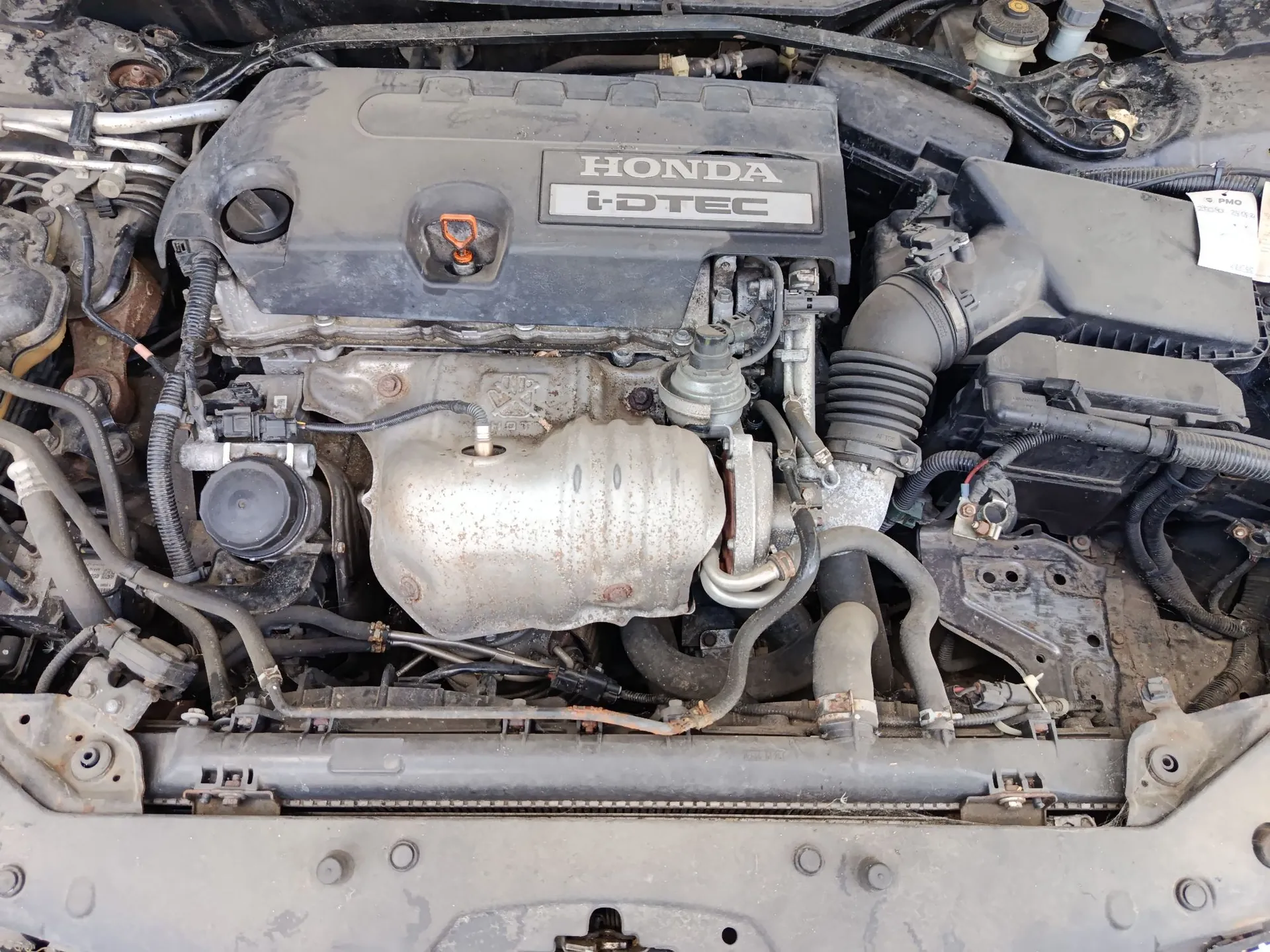 Widok z góry na komorę silnika samochodu marki Honda. Centralnie znajduje się plastikowa osłona silnika z napisem 'HONDA i-DTEC'. Widoczne są elementy mechaniczne, takie jak rury dolotowe, przewody, akumulator po prawej stronie oraz chłodnica na dole obrazu.