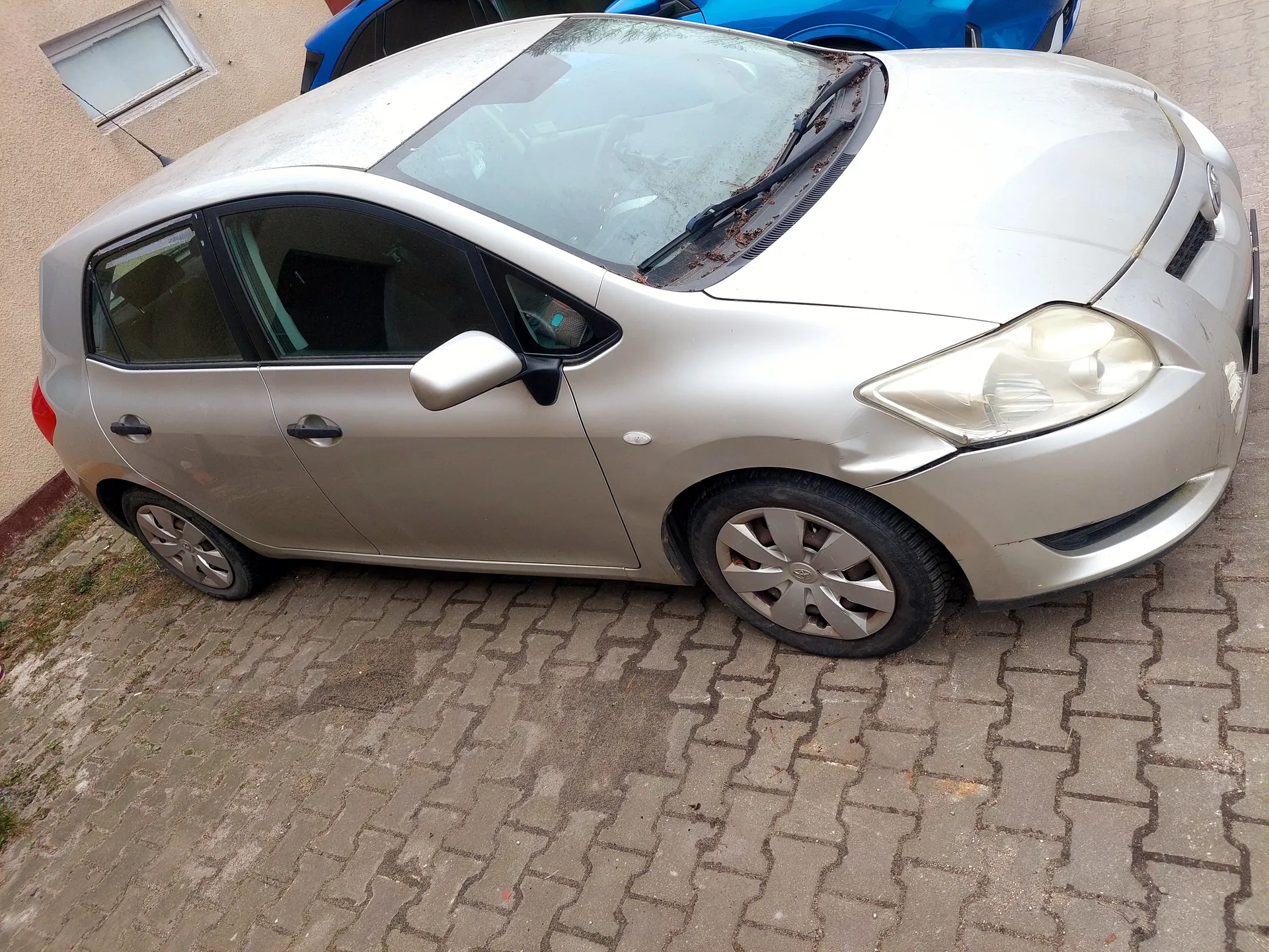 Srebrny samochód osobowy typu hatchback zaparkowany na brukowanej powierzchni. Widoczne uszkodzenie przedniej prawej części nadwozia oraz zanieczyszczenia na przedniej szybie. W tle niebieski samochód i fragment budynku z małym oknem.