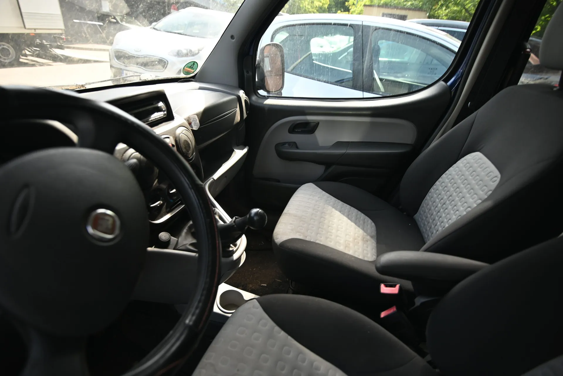 Wnętrze samochodu z perspektywy kierowcy. Widoczna jest czarna kierownica z logo marki Fiat, deska rozdzielcza, czarne fotele z jasnoszarymi, teksturowanymi wstawkami na siedziskach i oparciach. Przez otwarte okno pasażera widać zaparkowany biały samochód osobowy.