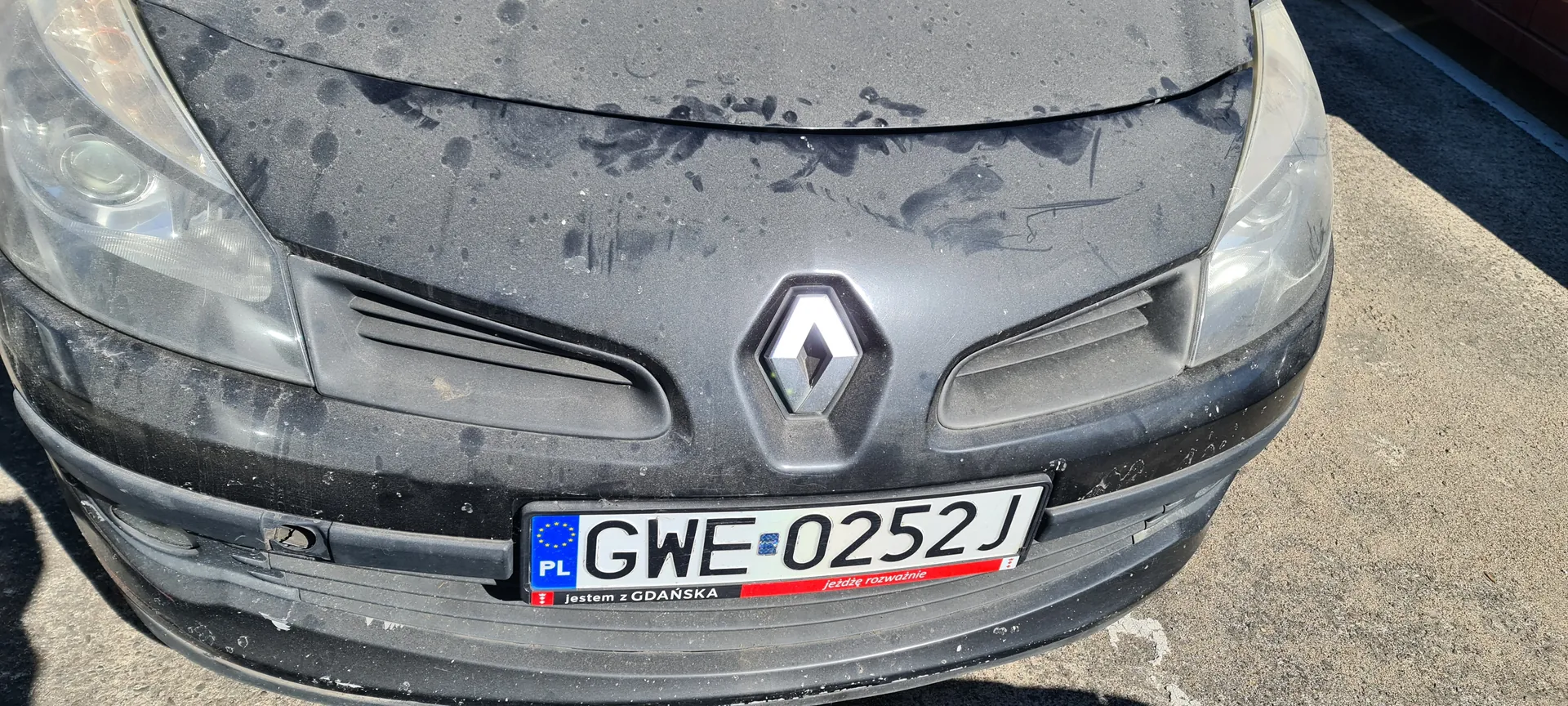 Przód czarnego samochodu marki Renault z widocznym logo w kształcie rombu na masce. Na masce znajdują się plamy i ślady brudu. Tablica rejestracyjna z białym tłem i czarnym napisem GWE-0252J, po lewej stronie niebieski pas z napisem PL i symbolem Unii Europejskiej. Poniżej tablicy czerwono-czarny naklejony napis: 'jestem z GDAŃSKA' oraz 'jeżdżę rozważnie'. Samochód stoi na asfaltowej nawierzchni.