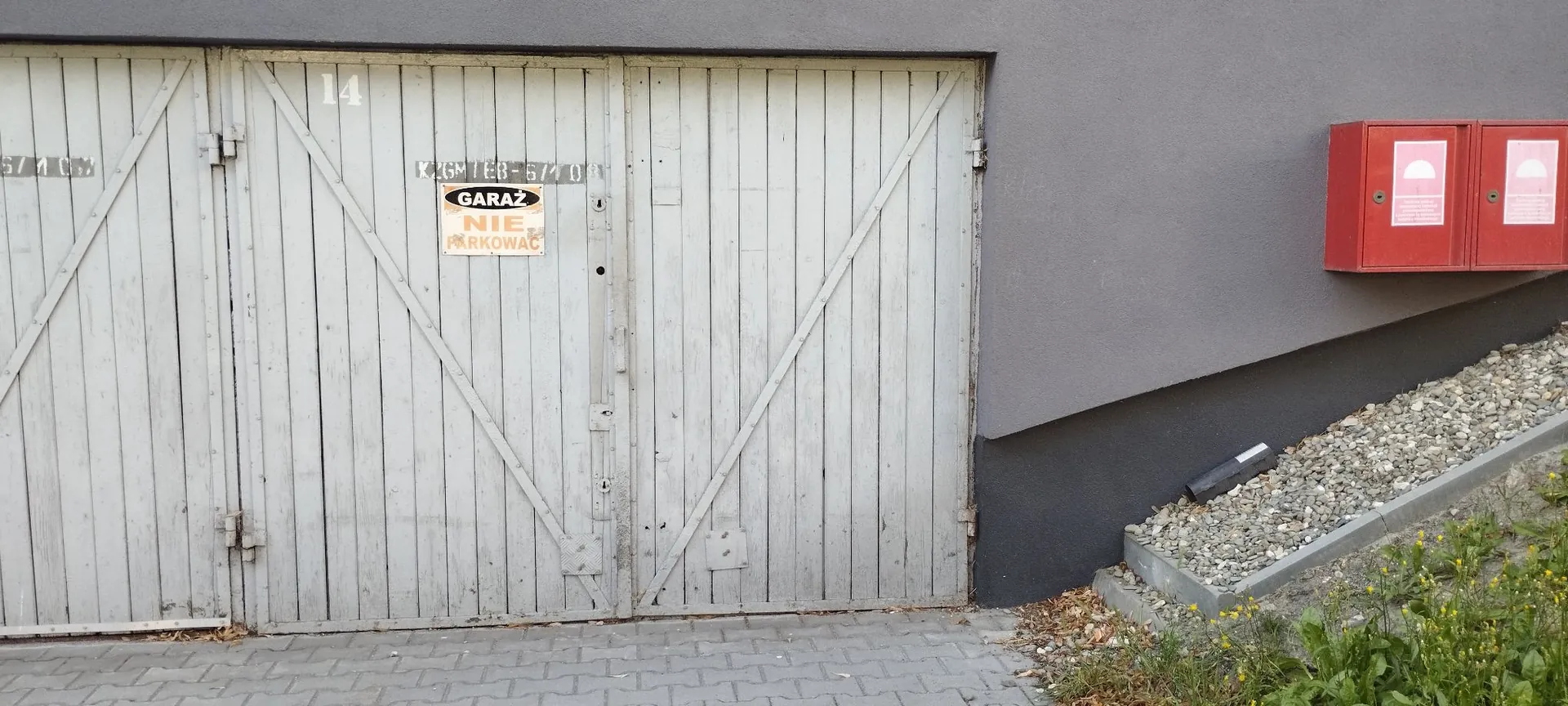 Zdjęcie przedstawiające białe, drewniane drzwi garażowe z pionowych desek, z widocznym numerem 14 nad nimi. Na drzwiach znajduje się tabliczka z napisem: 'GARAŻ NIE PARKOWAĆ'. Po prawej stronie znajduje się szara ściana budynku z zamontowanymi dwiema czerwonymi skrzynkami przeciwpożarowymi. Przed garażem widoczny jest chodnik z kostki brukowej, a obok ściany nasyp z szarego żwiru i fragmenty zieleni.