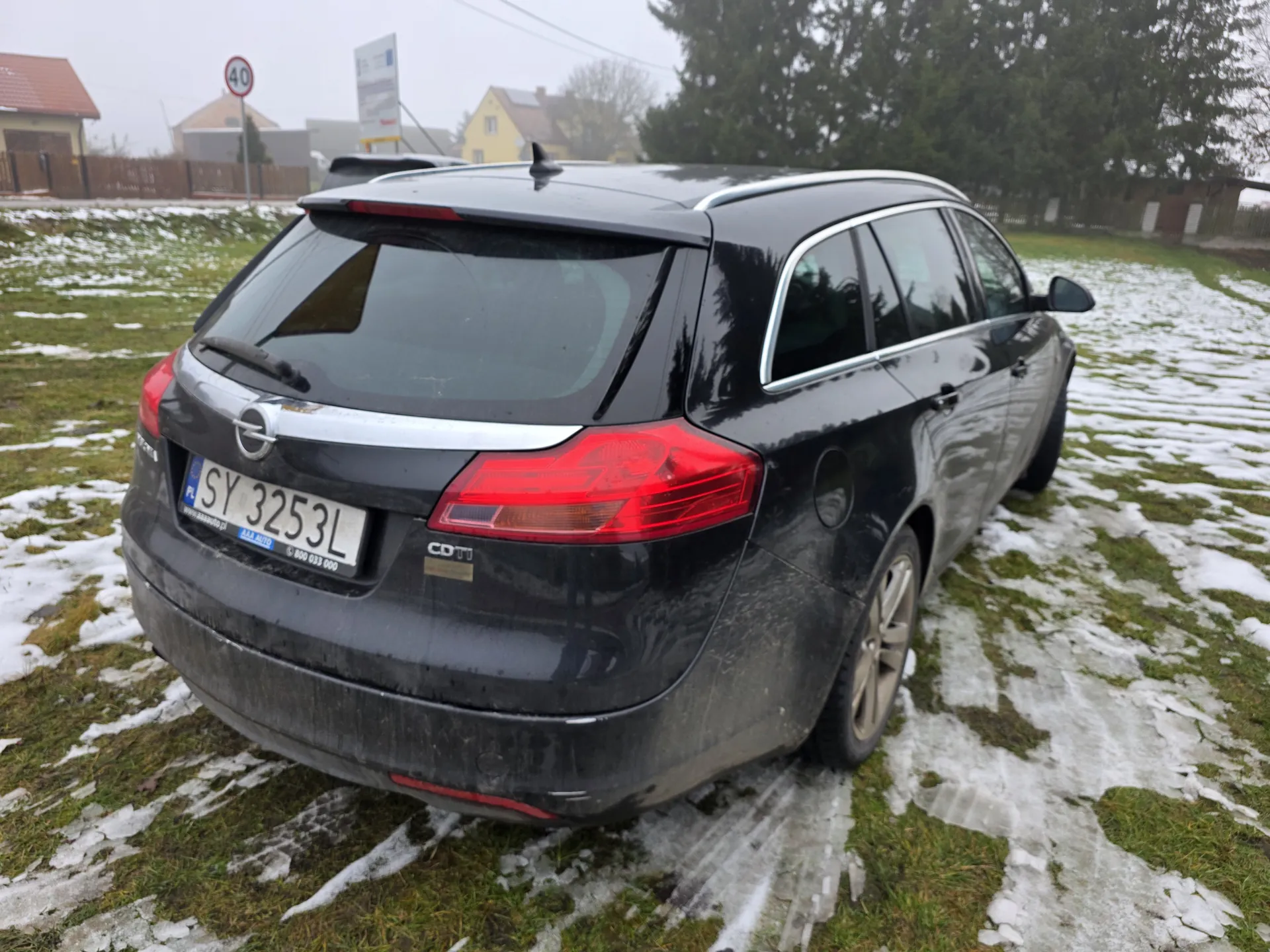 Czarny samochód osobowy typu kombi marki Opel z tablicą rejestracyjną SY 3253L, widok od tyłu. Samochód stoi na trawie z resztkami śniegu. W tle widoczne domy, znak drogowy 40, drzewa oraz mglisty krajobraz. Na tylnym zderzaku widoczny napis CDTi.
