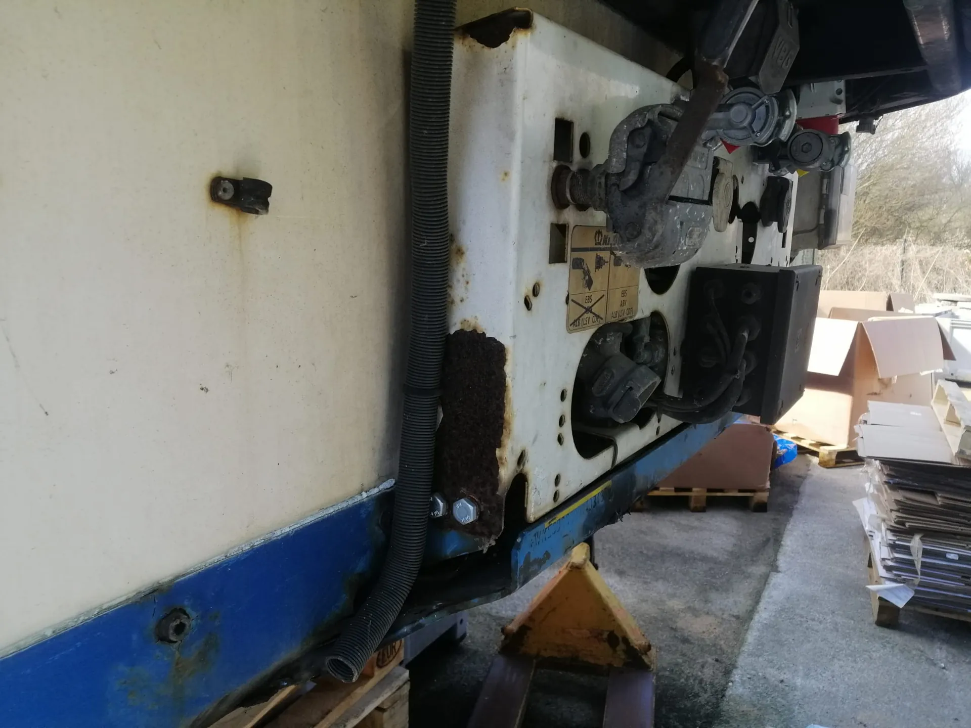 Zbliżenie na fragment białego nadwozia pojazdu z widocznymi elementami mechanicznymi, takimi jak metalowe wsporniki, śruby i czarny peszel prowadzący kable. Na białej powierzchni widoczne są ślady rdzy oraz mała żółta naklejka ostrzegawcza z piktogramami. Pod pojazdem znajduje się żółty element wspierający, a w tle widać betonowe podłoże i stosy kartonów.