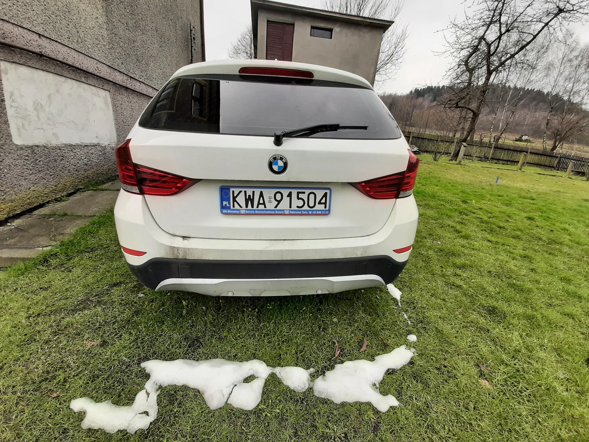 Biały samochód marki BMW widziany od tyłu, z tablicą rejestracyjną KWA 91504 z oznaczeniem PL po lewej stronie. Na trawie przed pojazdem znajdują się resztki śniegu. W tle budynki: po lewej betonowa ściana z białym plamą, za samochodem niewielki budynek z czerwonymi drzwiami i oknem. Po prawej stronie widoczny płot oraz drzewa bez liści. Niebo jest zachmurzone.