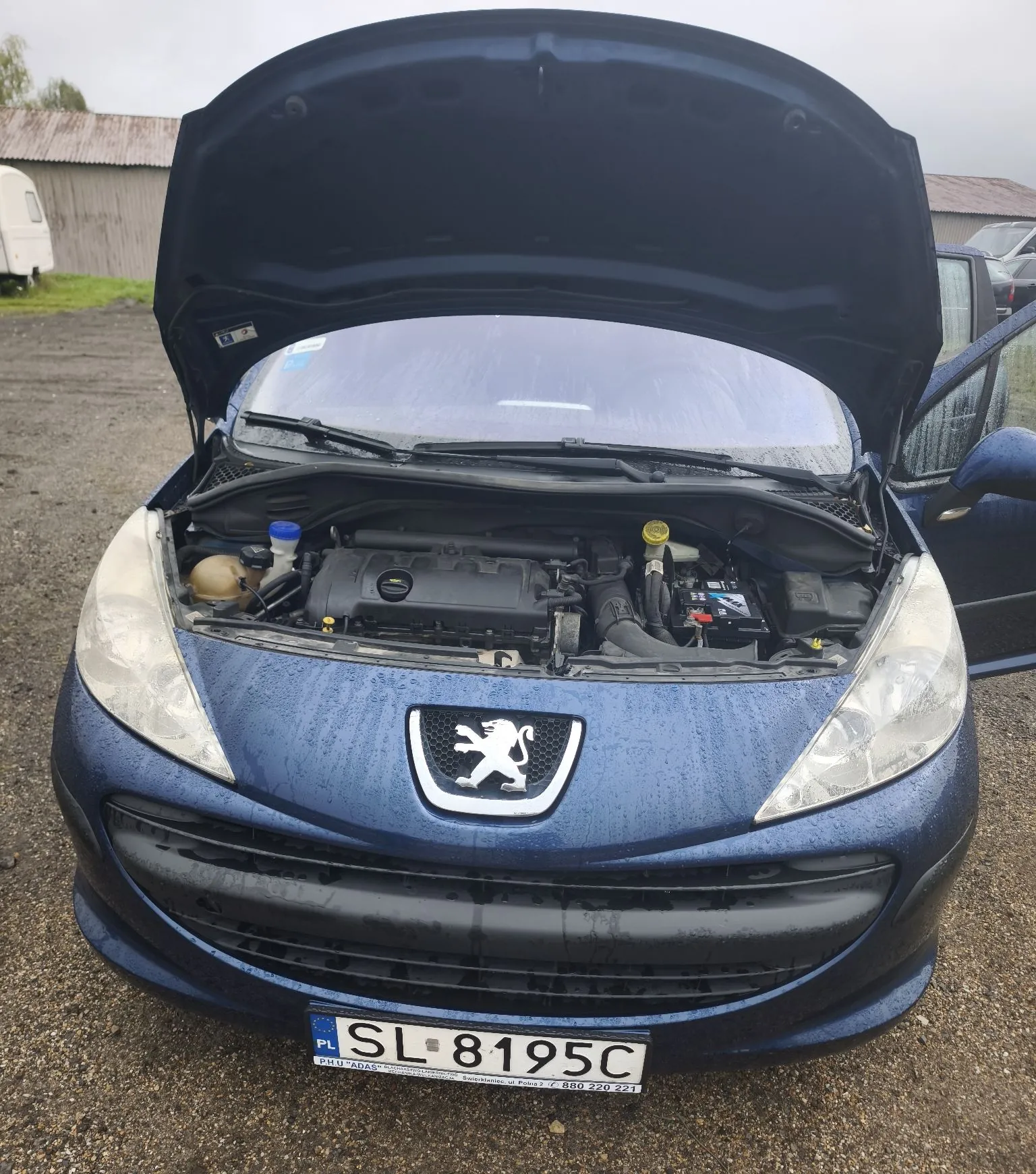 Niebieski samochód marki Peugeot z otwartą maską, ukazującą silnik. Pojazd stoi na żwirowym podłożu w deszczowy dzień, na karoserii widoczne są krople wody. Samochód posiada polską tablicę rejestracyjną o numerze SL 8195C.