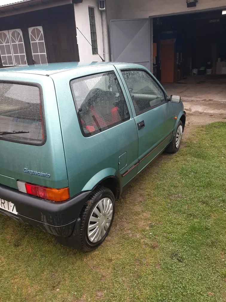 Zielony samochód Fiat Cinquecento zaparkowany na trawie przed budynkiem z otwartą bramą garażową. Widoczne są tylna część pojazdu z napisem "Cinquecento", tablica rejestracyjna z częściowo widocznym numerem, czerwone siedzenia wewnątrz oraz budynek z oknami i drzwiami w tle. Trawa na podwórku jest zielona, a na szybach samochodu znajdują się krople wody.