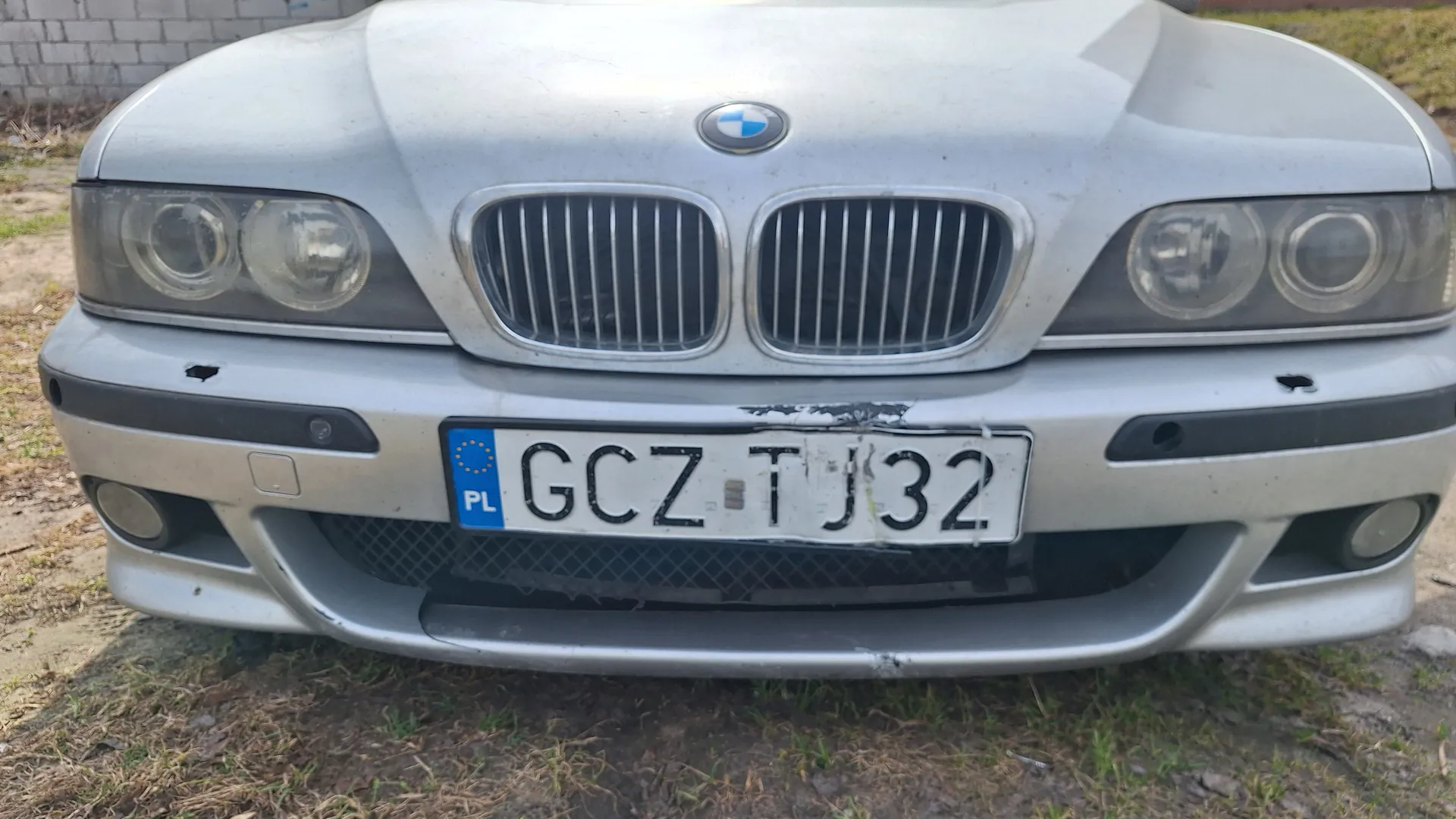 Przód srebrnego samochodu marki BMW z charakterystyczną podwójną atrapą chłodnicy, reflektorami i tablicą rejestracyjną. Tablica rejestracyjna zawiera napis GCZ T32 oraz niebieski element z oznaczeniem PL i symbolem Unii Europejskiej. Samochód posiada widoczne uszkodzenia na zderzaku. Tło stanowi trawa i bruk.