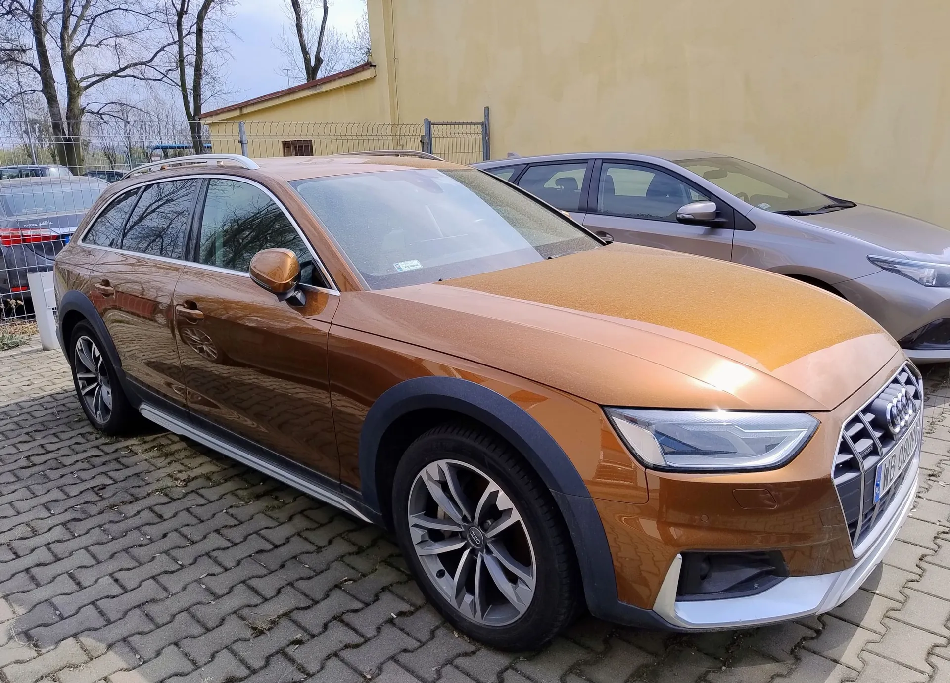 Pomarańczowy samochód marki Audi w nadwoziu kombi, zaparkowany na kostce brukowej. Auto jest widoczne z przodu i z boku. W tle znajduje się inny, szary samochód oraz żółta ściana budynku i bezlistne drzewa.