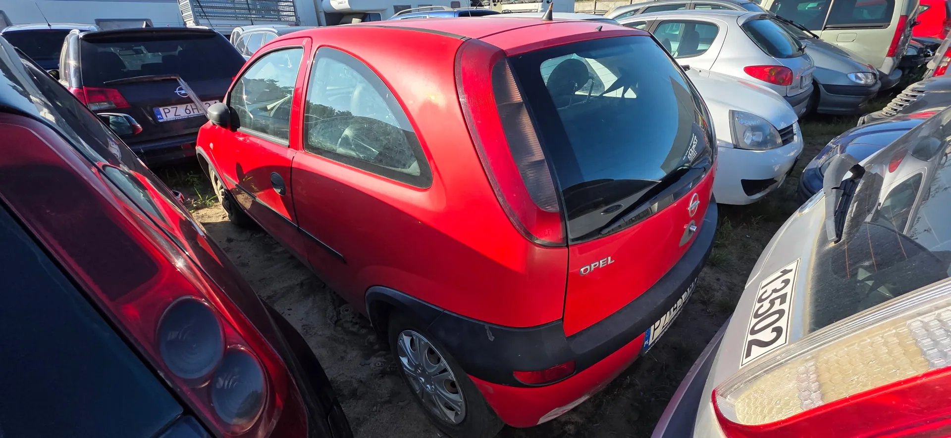Widok z tyłu i z boku czerwonego samochodu marki Opel Corsa zaparkowanego na placu wśród innych pojazdów. Samochód ma czarne plastikowe zderzaki i listwy. W tle widoczne są inne samochody w kolorach srebrnym, białym i czarnym. Na jednym z czarnych aut widoczna jest fragment tablicy rejestracyjnej PZ 6H.