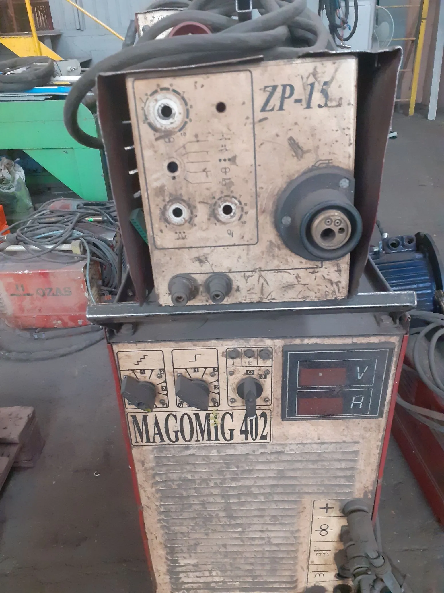 Zdjęcie starej, przemysłowej spawarki elektrycznej. Urządzenie składa się z dwóch głównych sekcji. Górna część to metalowa skrzynka z oznaczeniem 'ZP-15', posiadająca gniazda przyłączeniowe i pokrętła. Dolna część to obudowa z napisem 'MAGOMIG 402', wyposażona w trzy pokrętła regulacyjne, cyfrowy wyświetlacz z oznaczeniami 'V' (napięcie) i 'A' (natężenie prądu) oraz panel z symbolami elektrycznymi na dole. Urządzenie jest zakurzone i nosi ślady użytkowania.