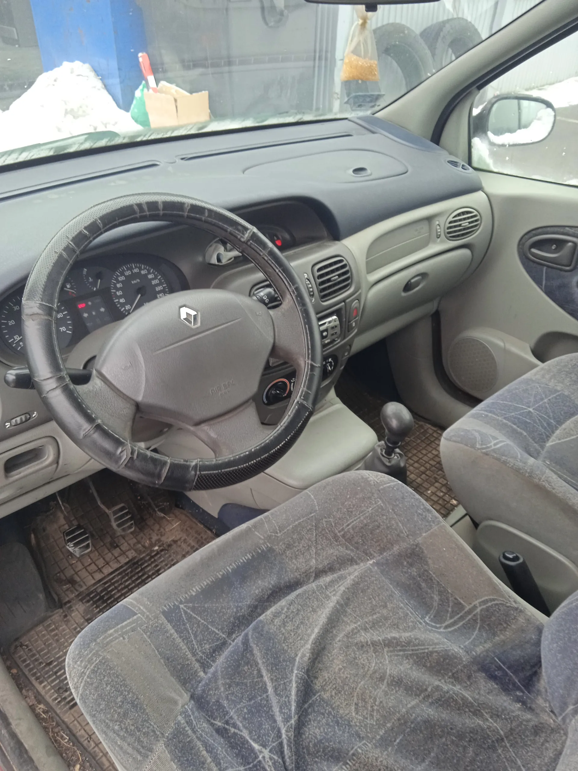 Wnętrze samochodu z widocznym kierownicą z logo, tablicą rozdzielczą z licznikami prędkości i obrotomierzem, szarymi siedzeniami z wzorem, brudnymi dywanikami podłogowymi oraz widokiem przez przednią szybę na zewnątrz, gdzie znajdują się opony i śnieg. Kierownica znajduje się po lewej stronie, a deska rozdzielcza jest w kolorze szaro-beżowym z wentylacjami i przyciskami.