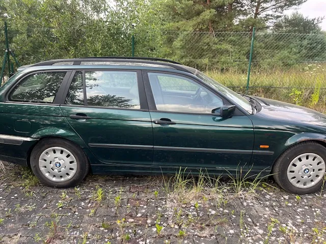 Boczny widok ciemnozielonego samochodu marki BMW w nadwoziu kombi, zaparkowanego na brukowanym podjeździe z wyrastającą trawą. W tle znajduje się zielony płot z siatki oraz gęste drzewa i krzewy.