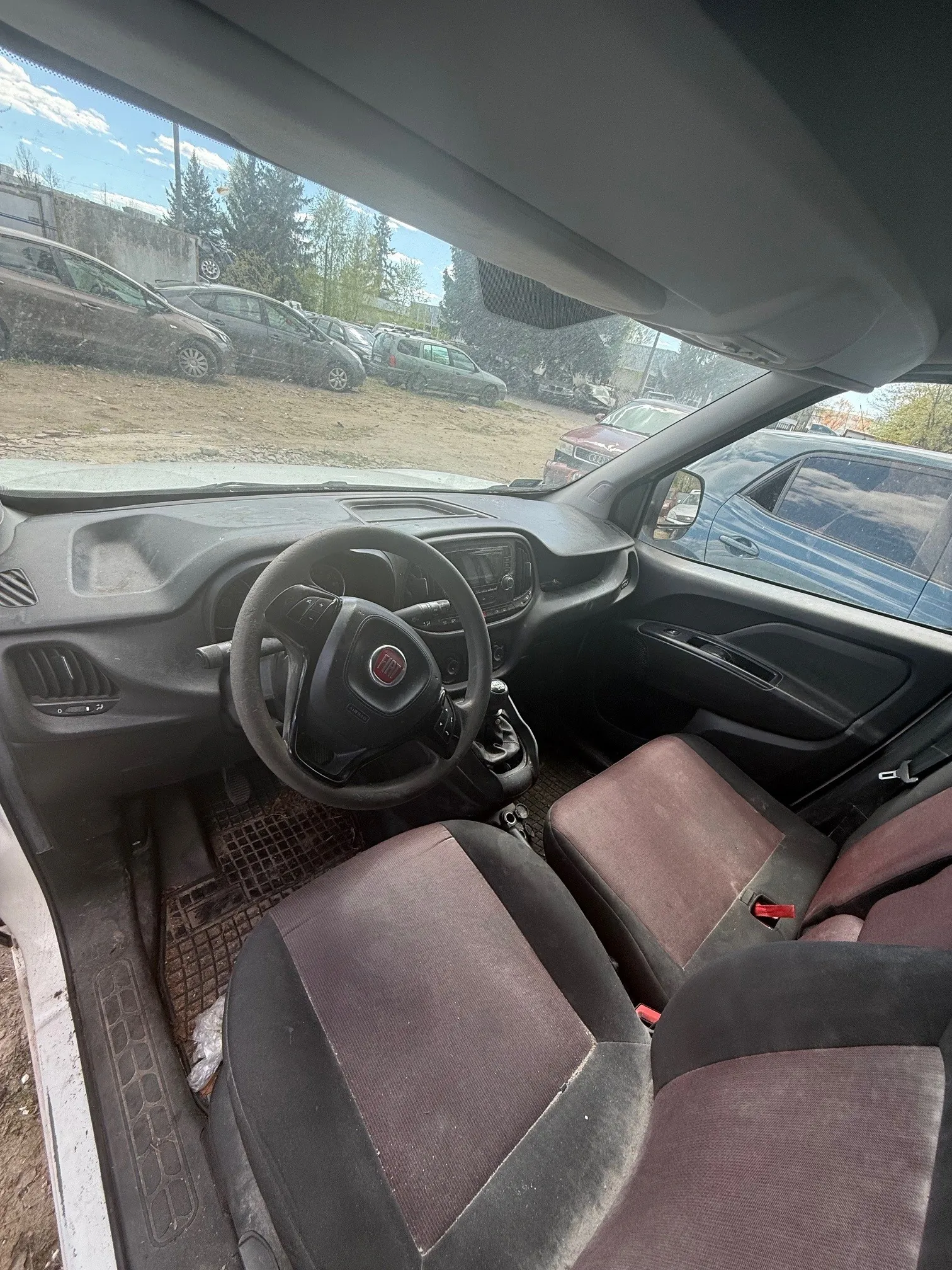 Wnętrze samochodu marki Fiat z widocznym kierownicą z czerwonym logo, czarno-brązowymi siedzeniami i szarą deską rozdzielczą. Przez przednią szybę widać parking z innymi pojazdami, drzewa i niebo z chmurami. Podłoga pokryta jest brudnymi dywanikami, a po lewej stronie widoczna jest otwarta drzwi samochodu.