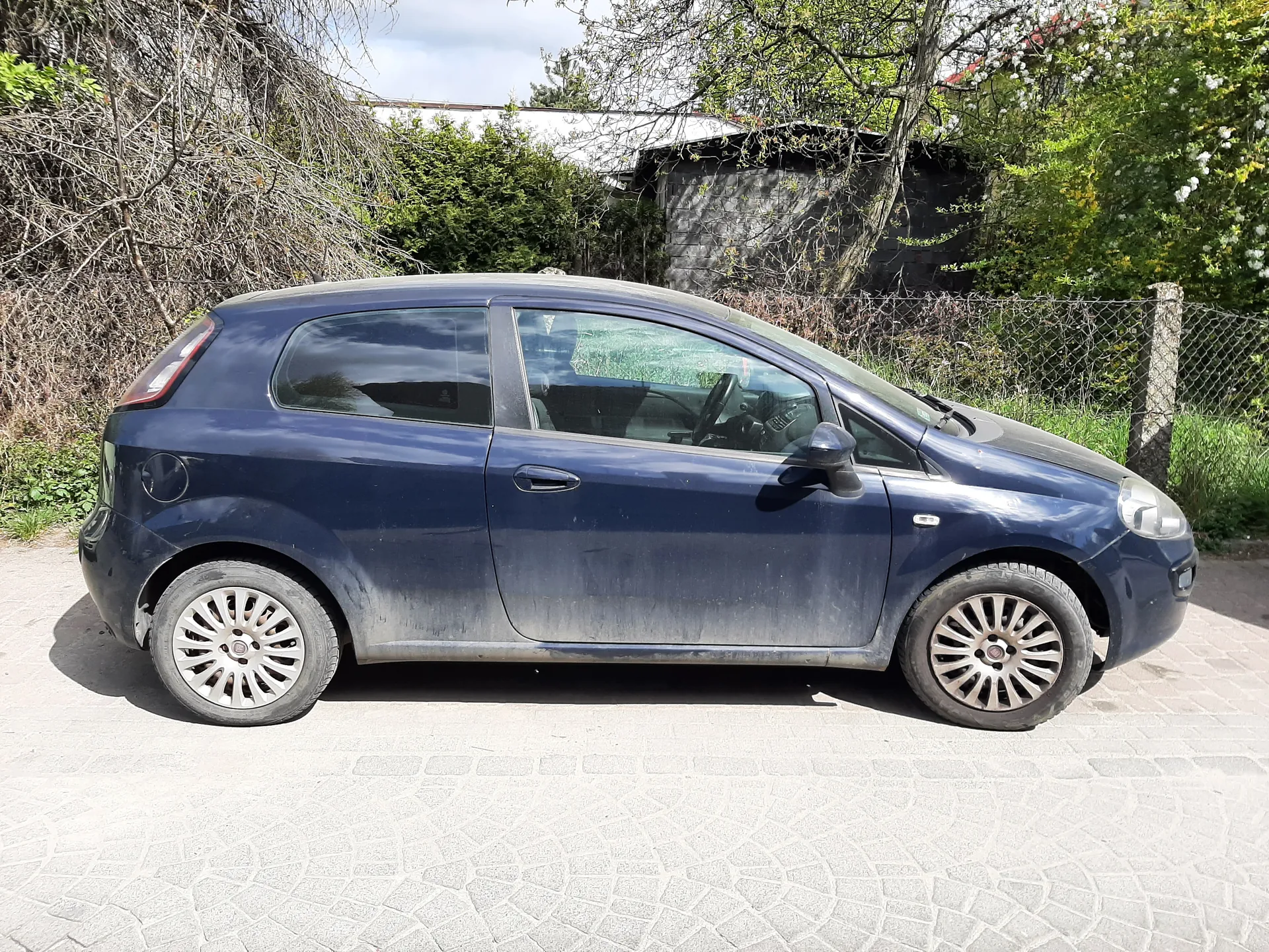 Niebieski samochód osobowy typu hatchback zaparkowany na brukowanej powierzchni. Pojazd jest zabrudzony, zwłaszcza dolne części nadwozia. W tle widoczne są zielone krzewy, drzewa, siatka ogrodzeniowa oraz kamienny budynek. Niebo jest częściowo zachmurzone.