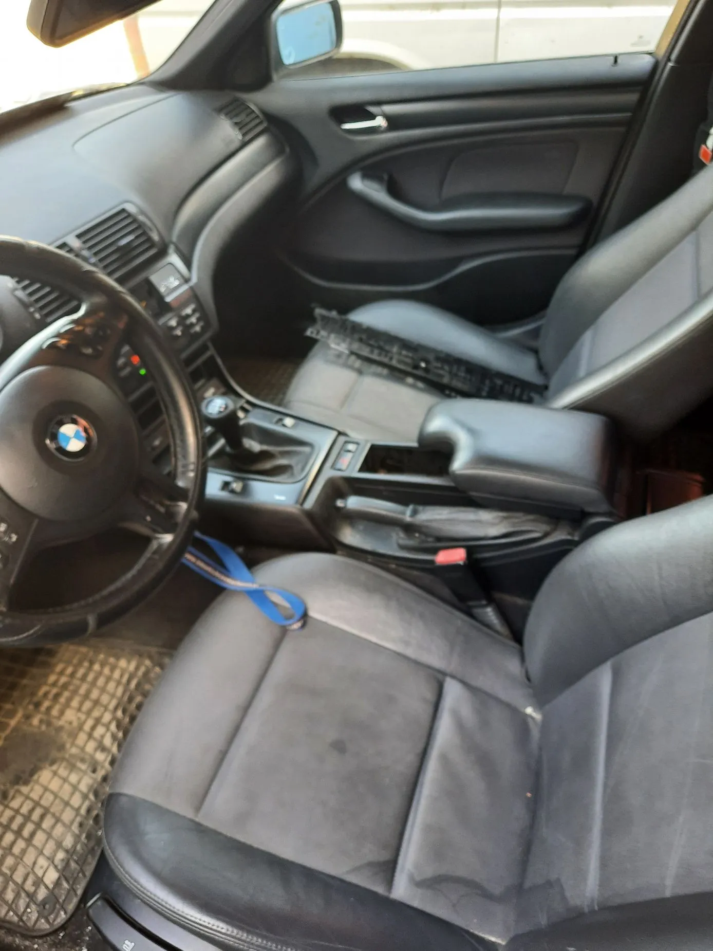 Wnętrze samochodu marki BMW z perspektywy kierowcy. Widoczna jest czarna skórzana tapicerka foteli, kierownica z logo BMW, konsola środkowa z lewarkiem zmiany biegów oraz przedni pasażerski fotel. Na podłodze znajduje się gumowa mata, a na fotelu kierowcy leży niebieska smycz.