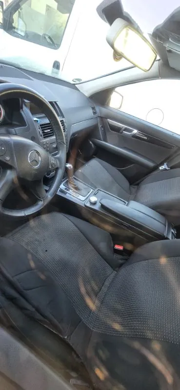 Wnętrze samochodu marki Mercedes-Benz z widocznym kierownicą z logo, czarnymi siedzeniami o strukturalnym wzorze, centralną konsolą z przyciskami i dźwignią zmiany biegów. Widoczne są również lusterko boczne, deska rozdzielcza oraz fragment przedniej szyby z odbiciem innego pojazdu. Wnętrze jest w kolorze ciemnoszarym, a na siedzeniach widoczne są ślady użytkowania.