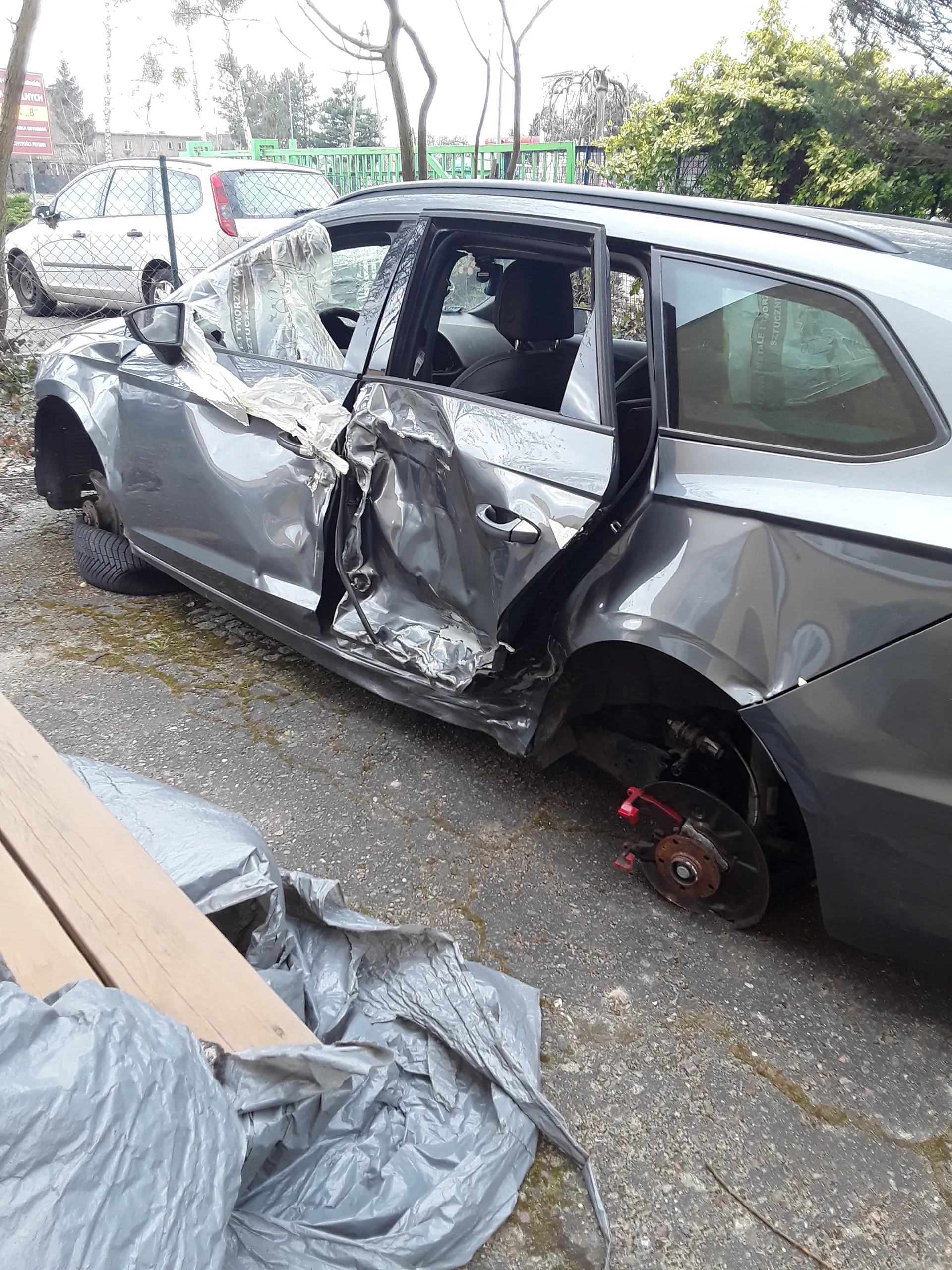 Zniszczony szary samochód osobowy z uszkodzoną przodem i lewą stroną. Brakuje przedniego lewego koła, widoczna tarcza hamulcowa z czerwonym elementem. Drzwi pasażerskie są otwarte, a nadwozie jest mocno zdeformowane. Na ziemi leży szary płacht, a w tle widoczny jest biały samochód, zielone konstrukcje i drzewa.