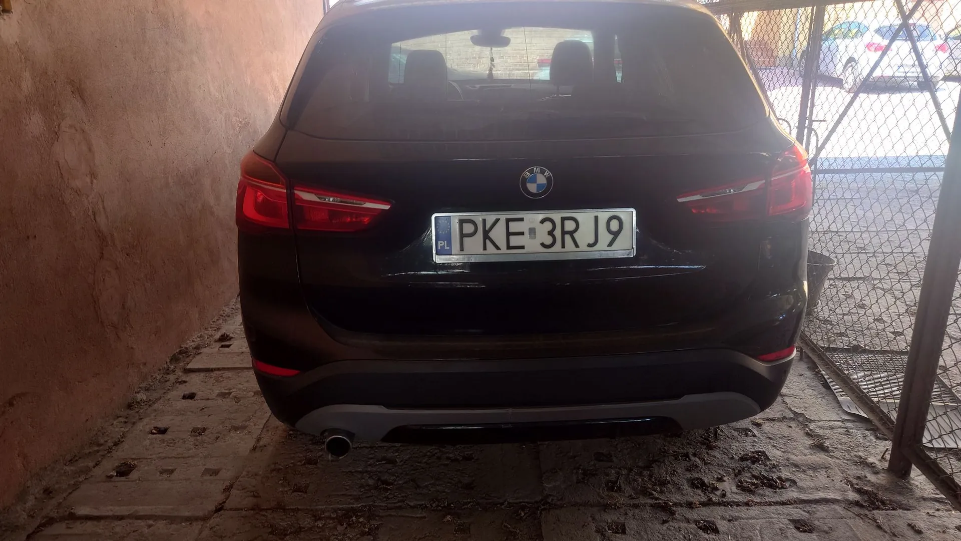 Tylny widok czarnego samochodu marki BMW zaparkowanego w garażu lub pod wiatą. Na samochodzie znajduje się polska tablica rejestracyjna o numerze PKE 3RJ9. Po lewej stronie widoczna jest brązowa ściana, a po prawej metalowa siatka ogrodzeniowa.