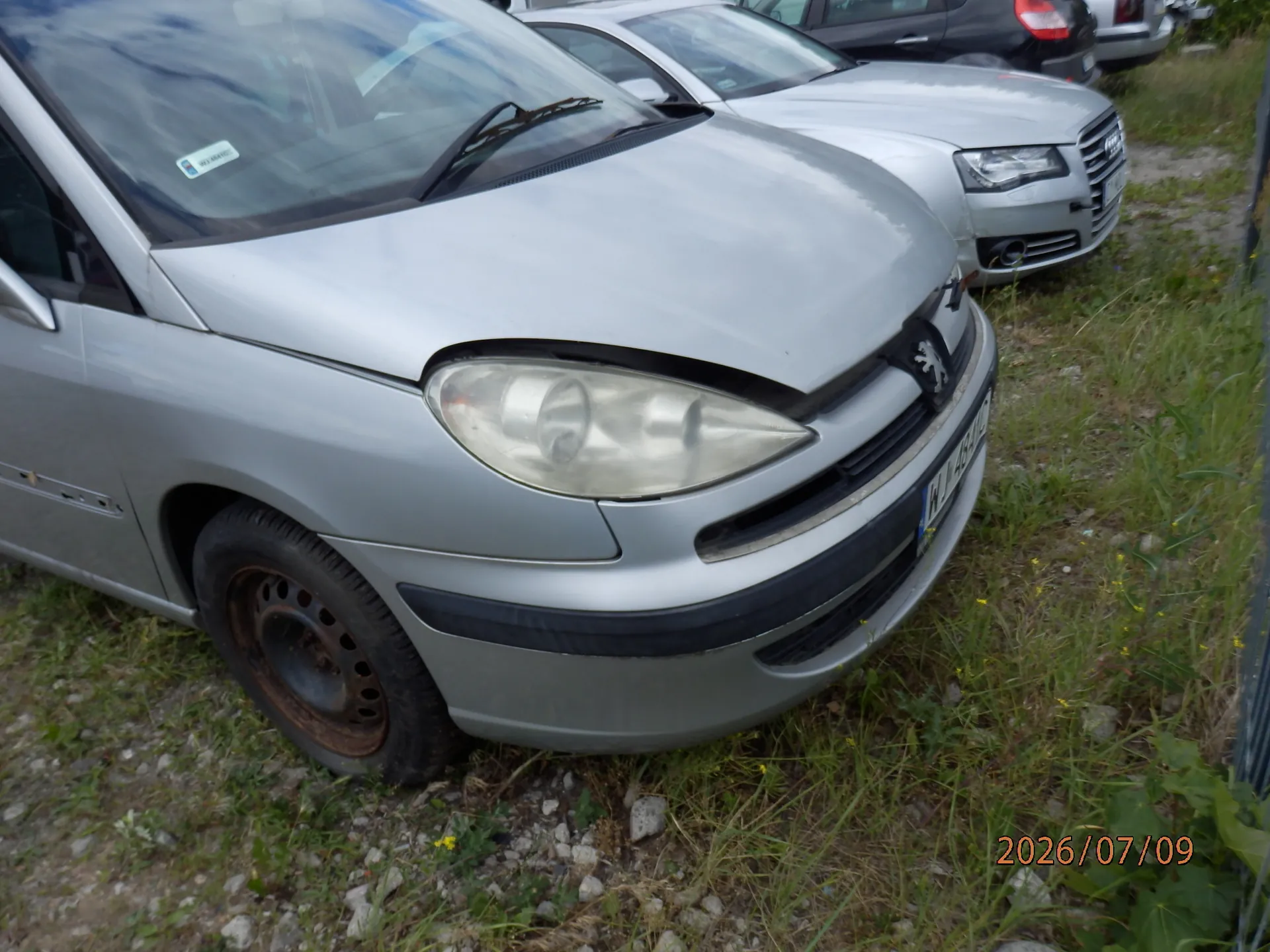 Srebrny samochód marki Peugeot zaparkowany na trawiastym terenie. Widoczny jest przód pojazdu z uszkodzoną maską, która jest lekko uniesiona nad reflektorem. Samochód ma polską tablicę rejestracyjną zaczynającą się od liter WK. W tle widoczne są inne zaparkowane samochody, w tym srebrne Audi.