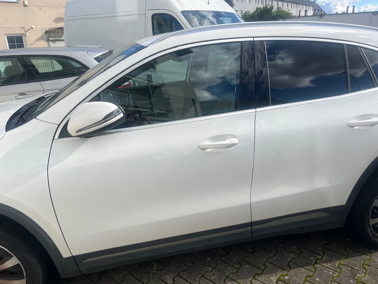 Zbliżenie na prawy bok białego samochodu typu SUV zaparkowanego na kostce brukowej. W tle widoczny jest fragment innego srebrnego samochodu oraz biały furgon.