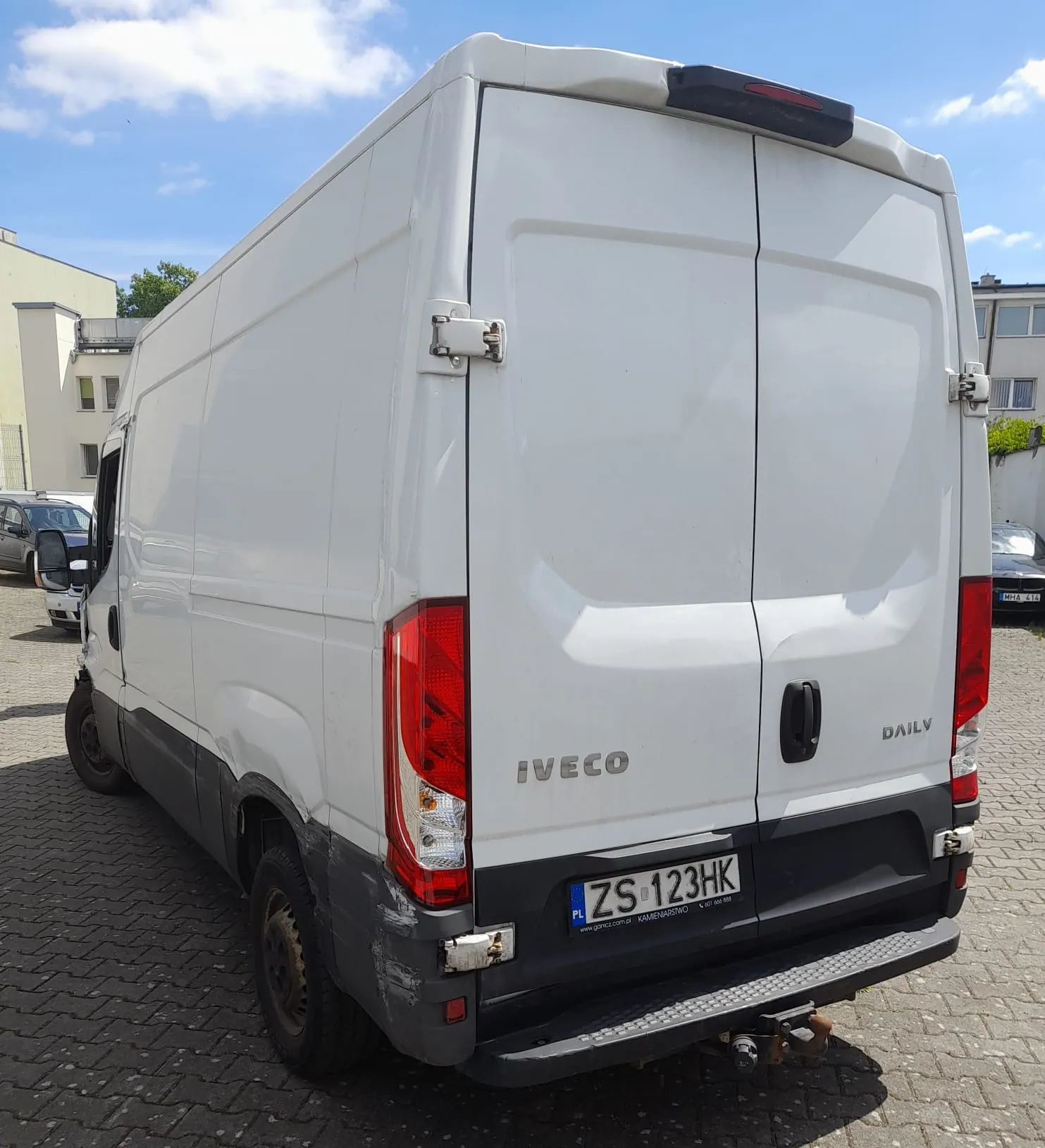 Biały furgon marki Iveco Daily zaparkowany na kostce brukowej. Zdjęcie przedstawia tył i lewy bok pojazdu. Na tylnych drzwiach widnieją napisy 'IVECO' oraz 'DAILY'. Tablica rejestracyjna pojazdu to ZS 123HK. Samochód posiada hak holowniczy oraz widoczne uszkodzenia lakieru na dolnej części lewego tylnego panelu.