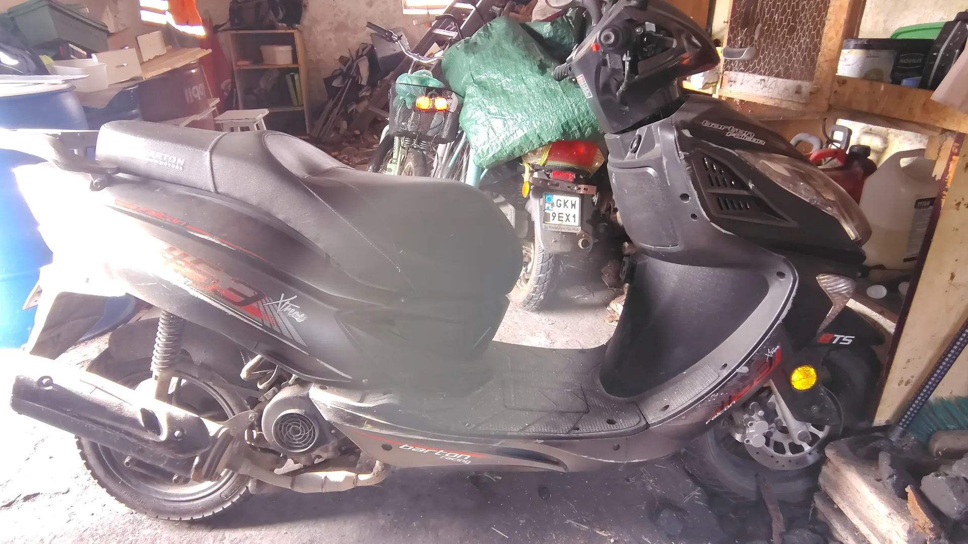 Czarny skuter marki Barton z napisem Xtreme i numerem 6TS na boku, umieszczony w przestronnym garażu. W tle widoczny drugi skuter z tablicą rejestracyjną GKW 9EX1, zielona folia, półki z narzędziami, pojemniki i różne przedmioty. Podłoga z betonu, ściany z widocznymi śladami zużycia.