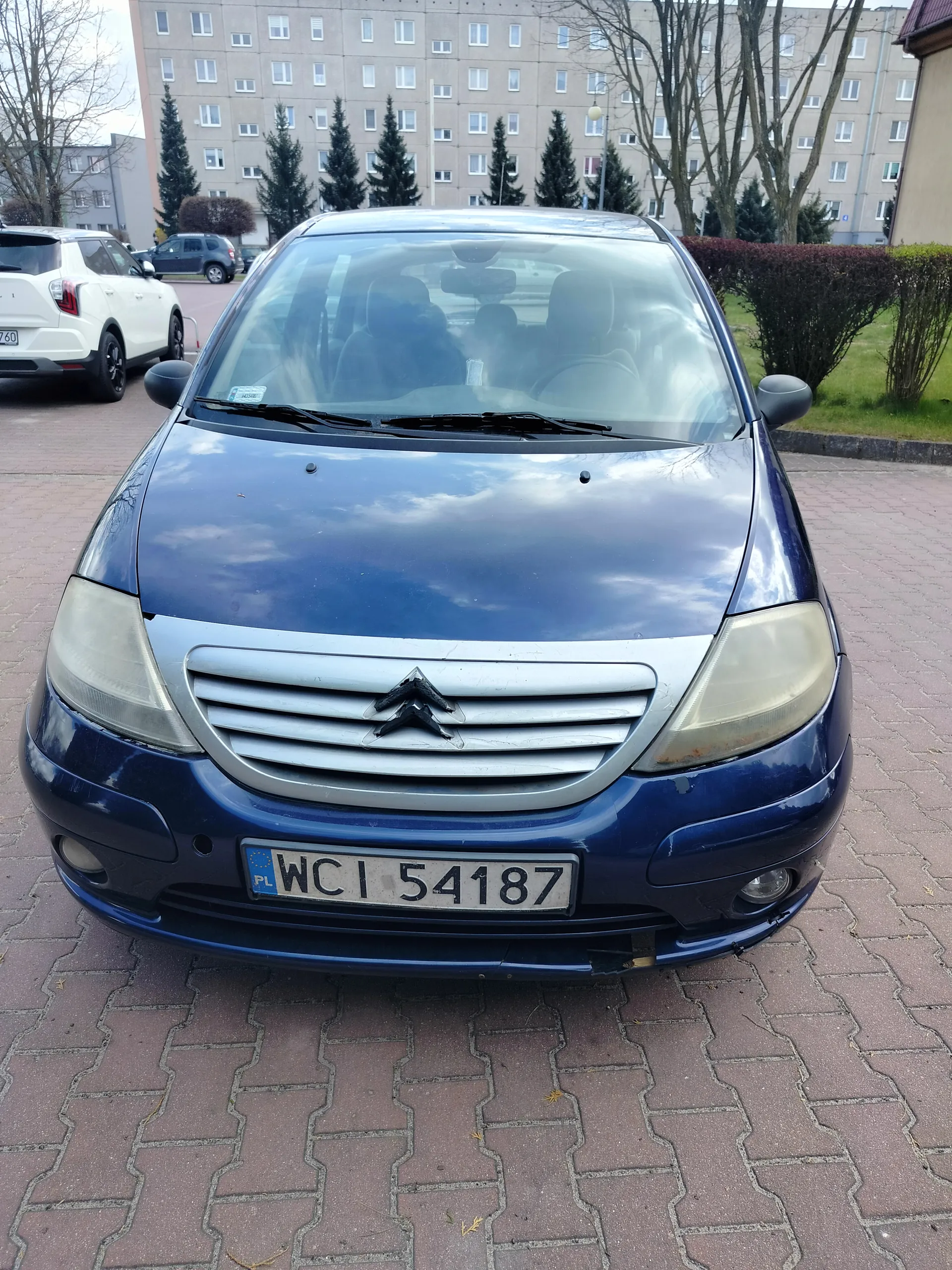 Niebieski samochód marki Citroën z tablicą rejestracyjną WCI 54187, zaparkowany na brukowanym placu. W tle budynek mieszkalny z oknami, drzewa, krzewy oraz biały samochód. Tablica rejestracyjna zawiera oznaczenie PL oraz numer WCI 54187.
