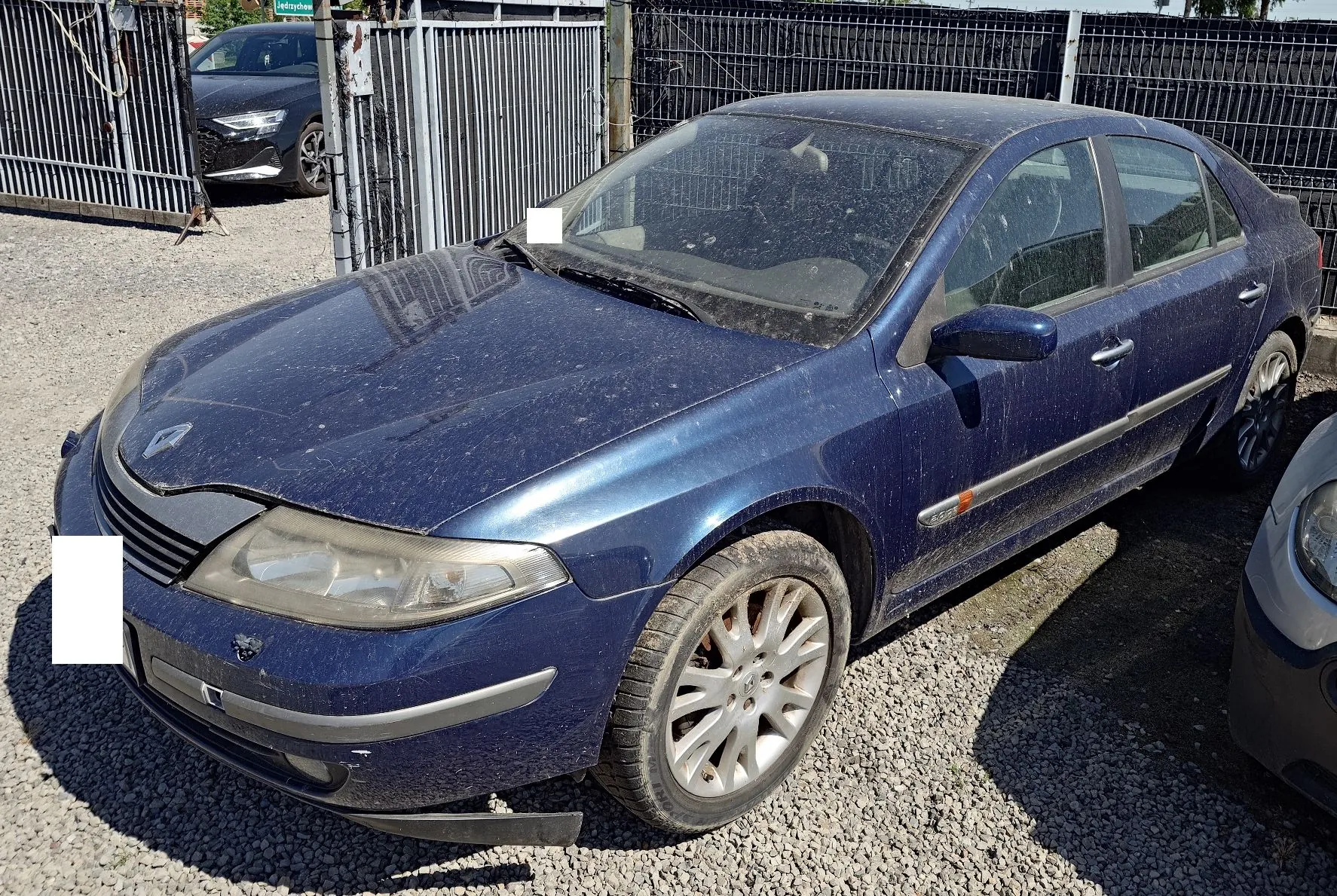 Niebieski samochód osobowy marki Renault Laguna zaparkowany na żwirowym placu. Pojazd jest zakurzony, z widocznymi zabrudzeniami na karoserii. Auto stoi pod kątem, widoczny jest przód oraz prawy bok. W tle znajduje się metalowy płot i fragment innego, ciemnego samochodu.