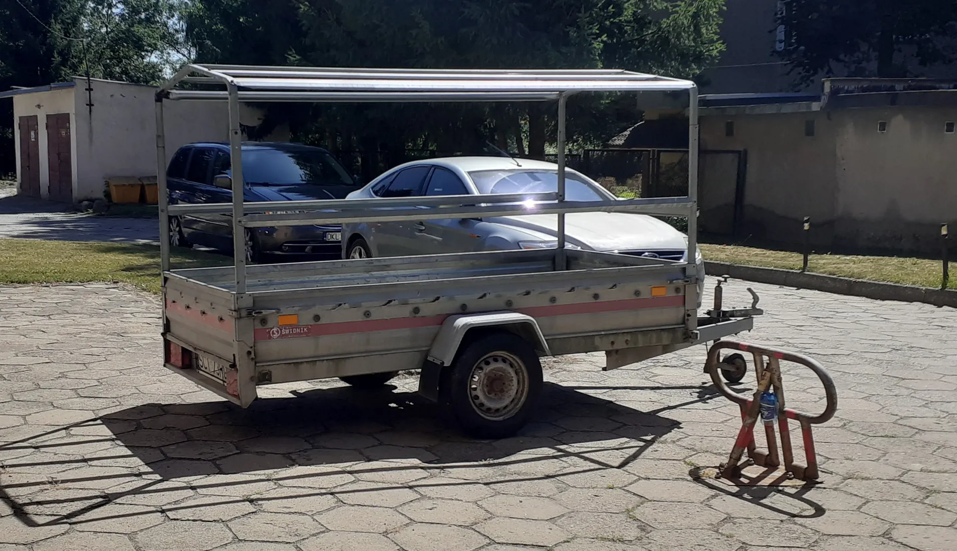 Srebrna przyczepa transportowa z metalową konstrukcją zadaszenia, zaparkowana na kostce brukowej. W tle widoczne są dwa samochody: ciemny minivan oraz srebrny sedan, a także budynki i drzewa.