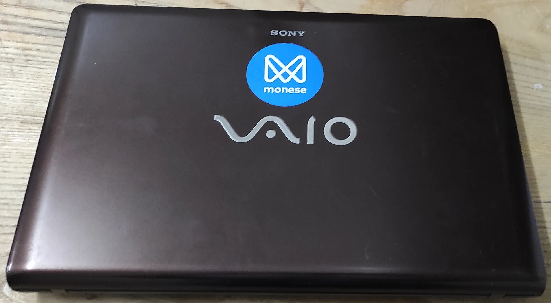 Zamknięta pokrywa ciemnobrązowego laptopa marki Sony VAIO. Na górze znajduje się biały napis SONY, poniżej niebieska okrągła naklejka z białym logo i napisem 'monese', a w centrum duży, srebrny napis VAIO.