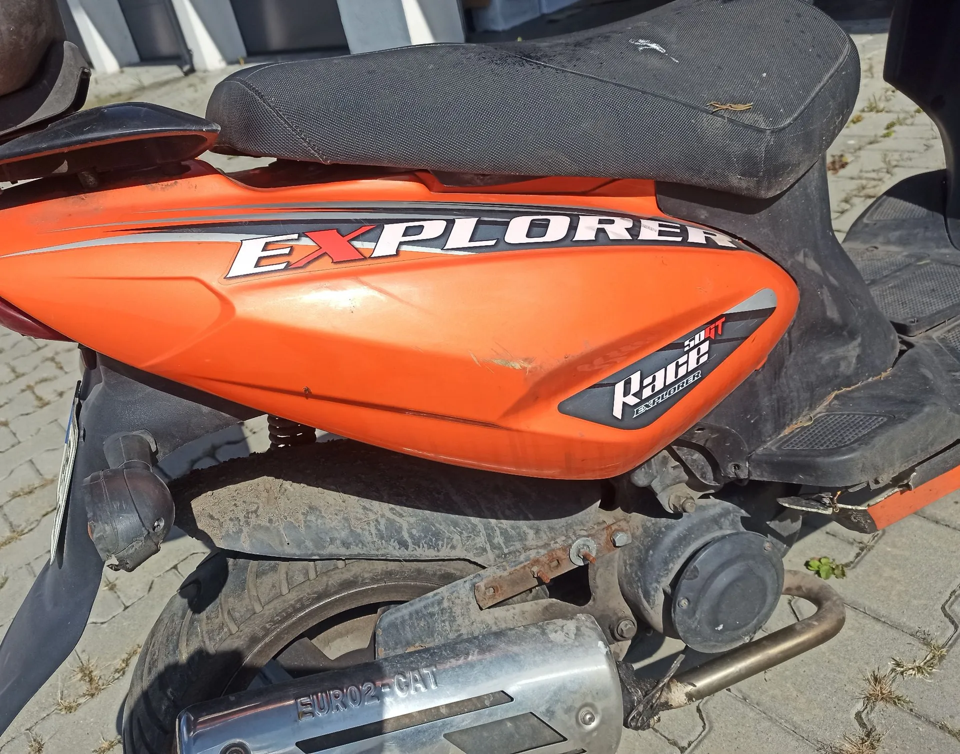 Zbliżenie na tylną część pomarańczowego skutera. Na boku obudowy znajduje się biały napis 'EXPLORER' oraz mniejszy napis 'Race EXPLORER'. Widoczny jest czarny siedzenie, tylne koło, rura wydechowa z napisem 'EURO2-CAT' oraz fragmenty plastikowych osłon.