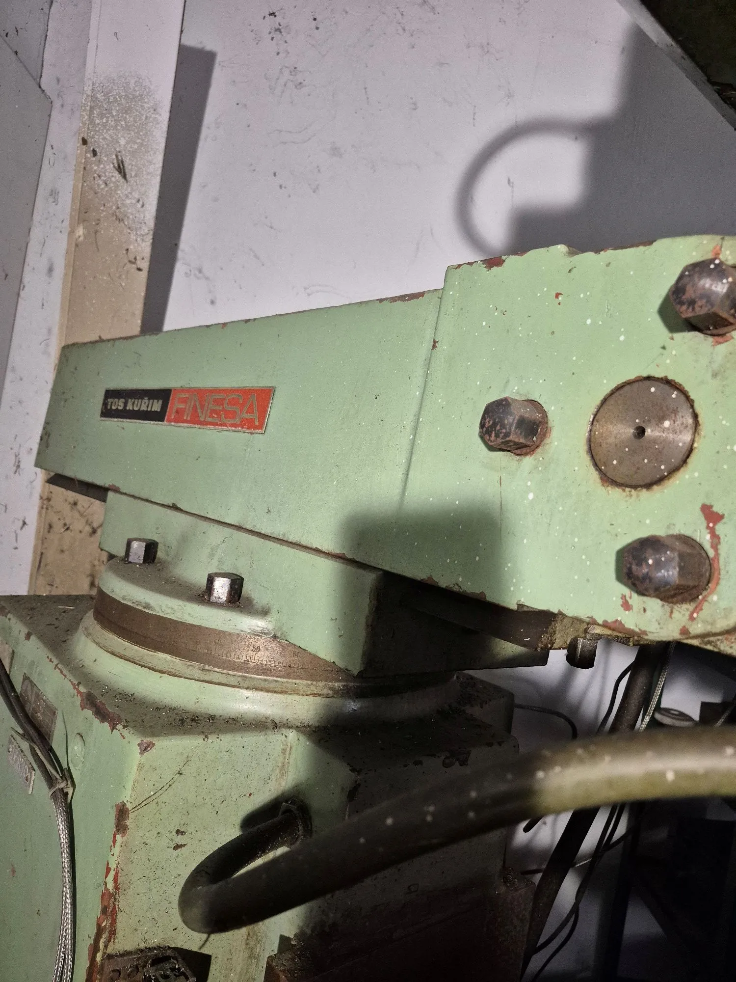 Zbliżenie na fragment zielonej, metalowej maszyny przemysłowej z widocznymi śladami zużycia i rdzy. Na boku urządzenia znajduje się czerwono-biała tabliczka z napisem „TOS KURIM FINESA”. Maszyna posiada duże śruby mocujące oraz metalowe elementy konstrukcyjne. W dolnej części widoczne są czarne przewody hydrauliczne lub elektryczne.