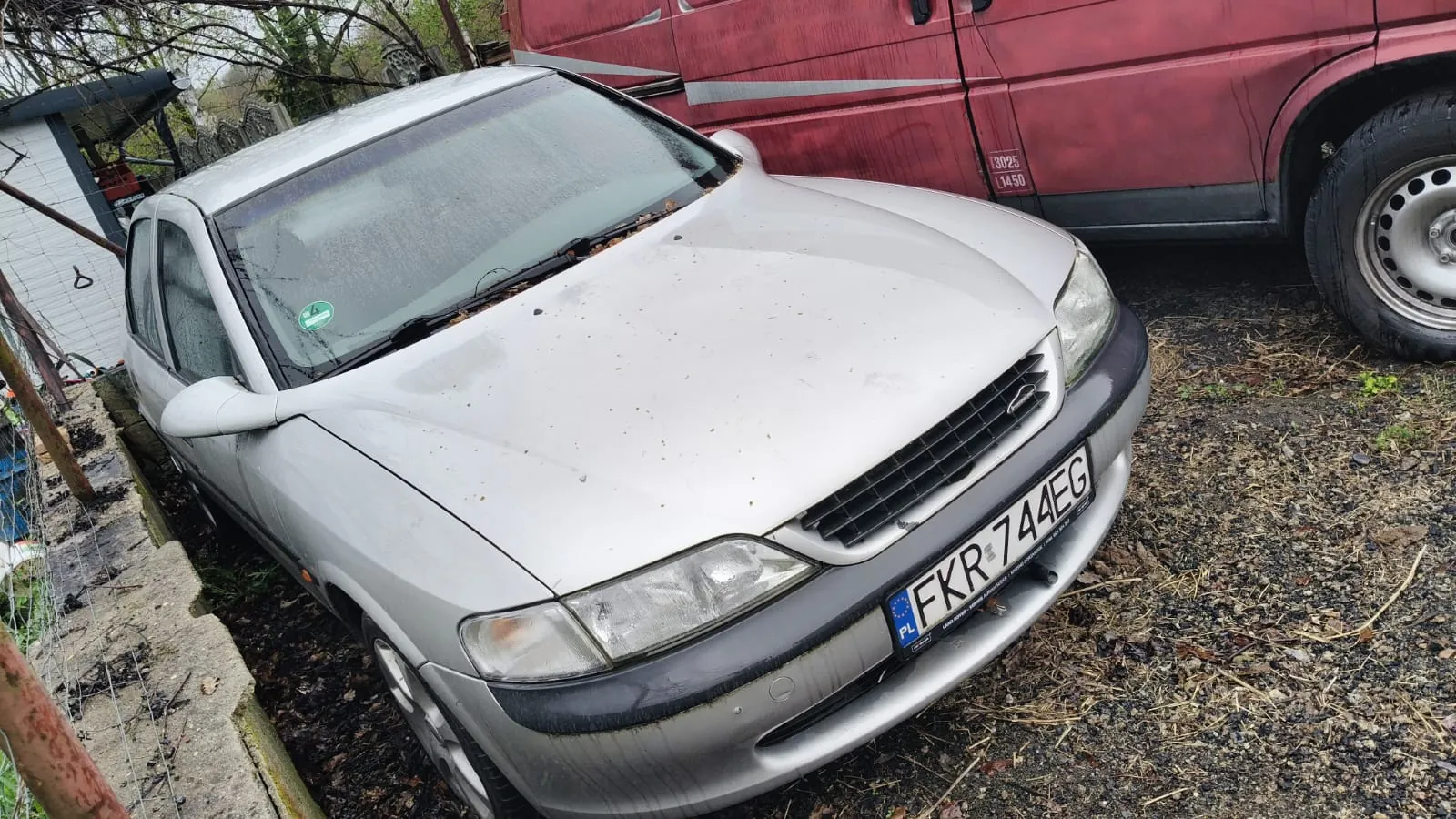 Srebrny samochód osobowy zaparkowany na podwórku z żwirem i suchymi liśćmi. Na przodzie widoczna tablica rejestracyjna PL FKR 744EG. Po prawej stronie czerwony pojazd dostawczy z napisami 3025 i 1450 na boku. W tle biały budynek, ogrodzenie i drzewa. Samochód ma zabrudzony nadwozie i zielony znaczek na przedniej szybie.