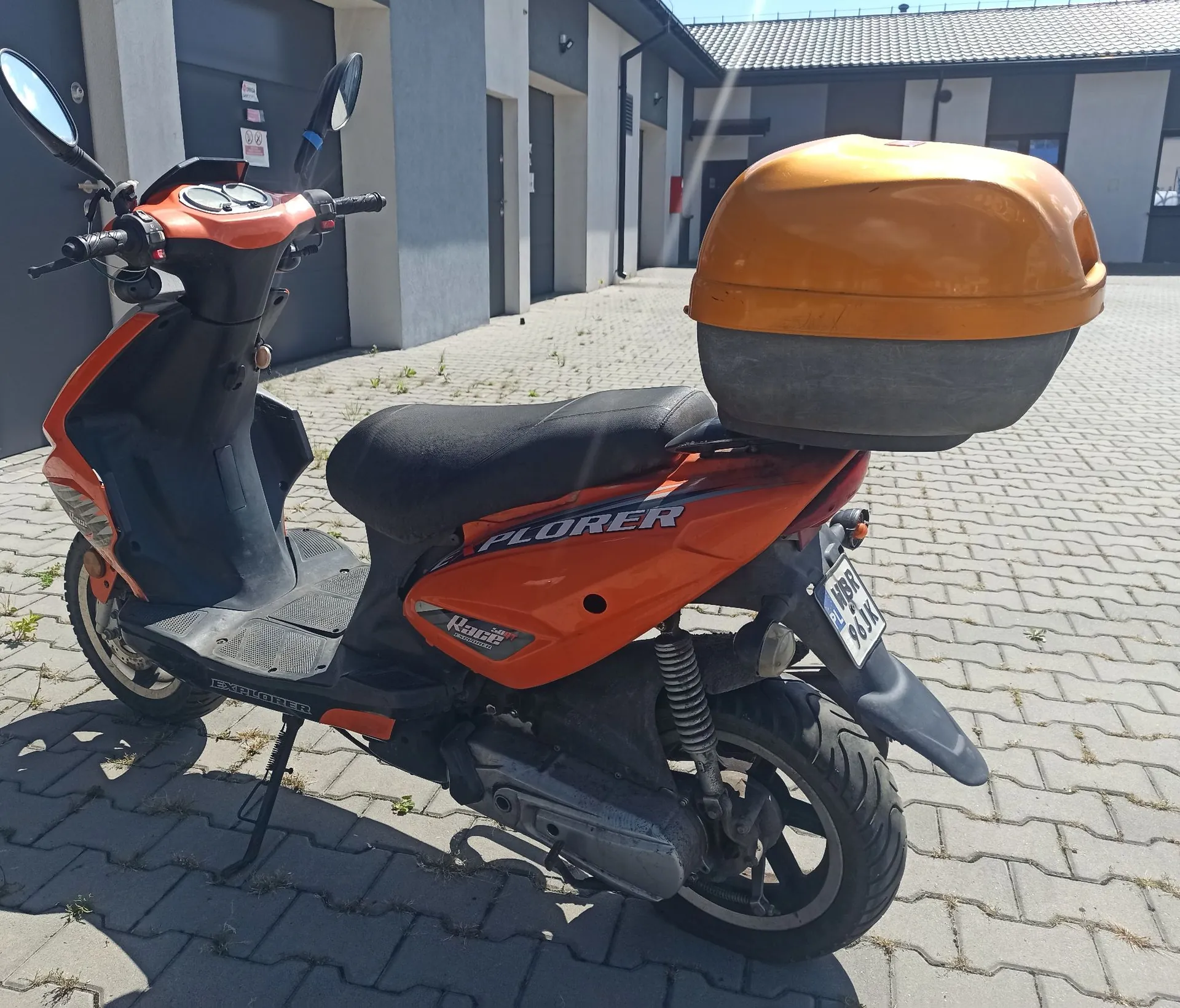 Pomarańczowy skuter marki Explorer zaparkowany na kostce brukowej przed szarym budynkiem. Skuter posiada czarną kanapę oraz zamontowany z tyłu pomarańczowy kufer transportowy. Widoczna jest tablica rejestracyjna oraz napis 'EXPLORER' na boku i podłodze pojazdu.