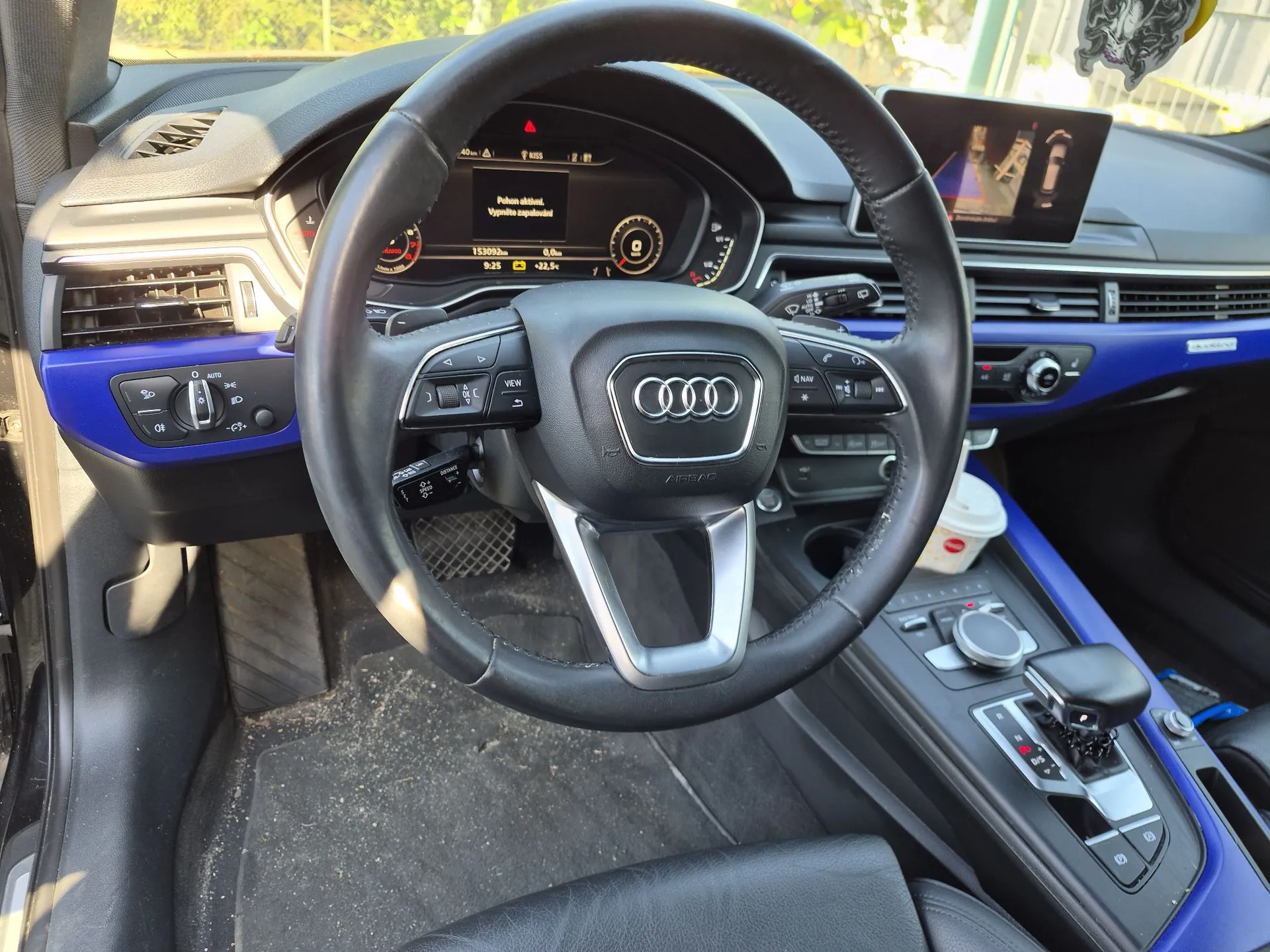 Wnętrze samochodu marki Audi z perspektywy kierowcy. Widoczna jest skórzana kierownica z logo Audi na środku, cyfrowy zestaw wskaźników z prędkościomierzem i obrotomierzem oraz ekran multimedialny po prawej stronie. Deska rozdzielcza posiada niebieskie akcenty dekoracyjne. W konsoli środkowej znajduje się lewarek zmiany biegów, pokrętło sterujące oraz kubek z napojem.