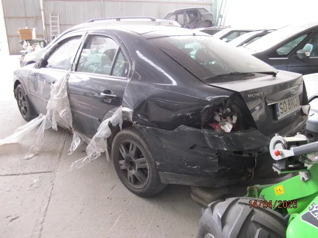 Czarny samochód osobowy marki Ford Mondeo z widocznym uszkodzeniem tylnego zderzaka i części karoserii. Na tylnym zderzaku widoczny napis 'MONDEO' oraz tablica rejestracyjna 'SO 8903B'. Część boczna pojazdu przykryta przezroczystą folią. W tle inne samochody, garaż oraz drabina. W prawym dolnym rogu obrazu widoczna data '14/04/2025' oraz fragment zielonego pojazdu lub maszyny. Podłoga betonowa, otoczenie przypomina warsztat lub stację naprawczą.