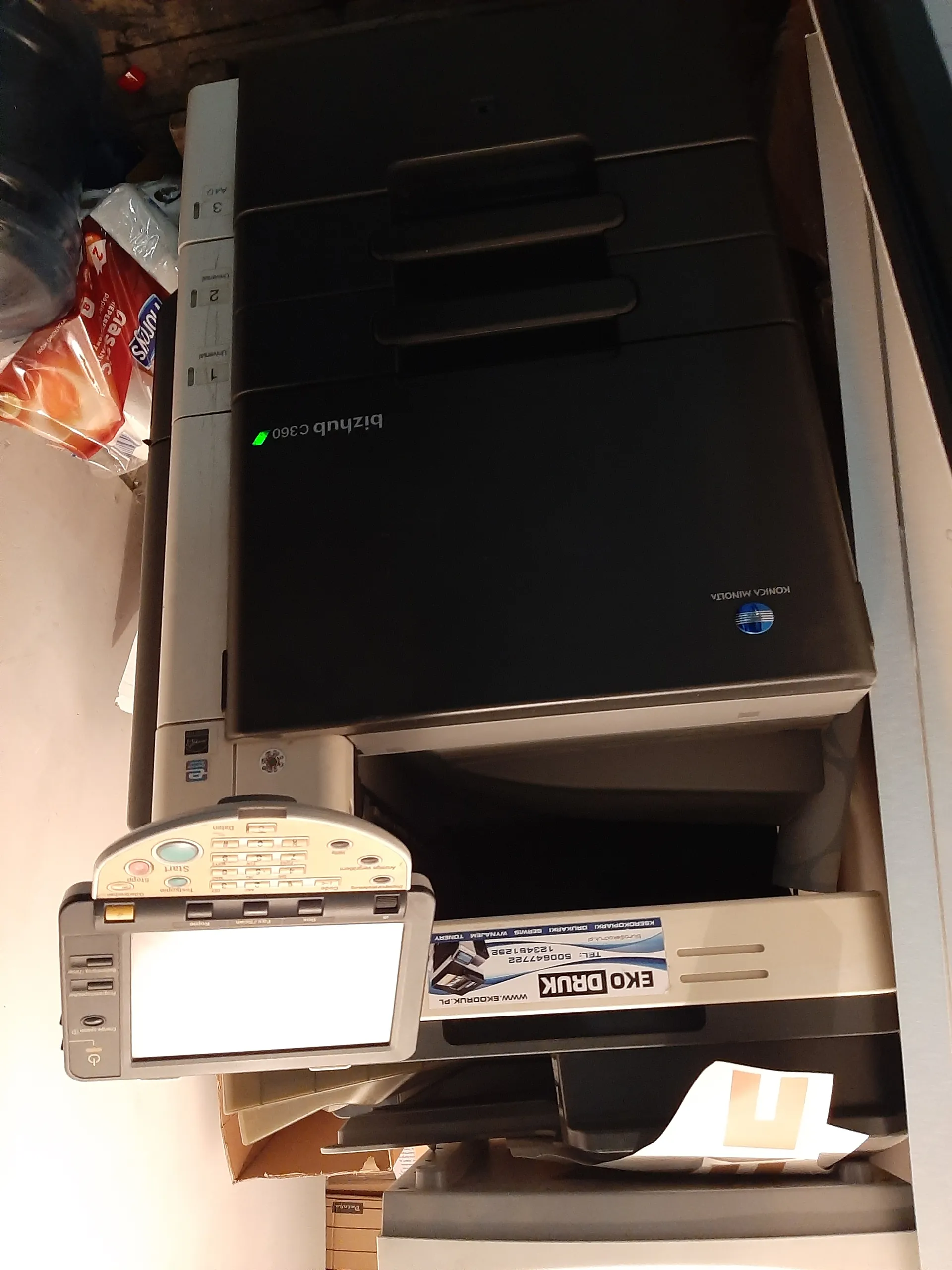 Drukarka wielofunkcyjna Konica Minolta bizhub c360 w kolorze czarno-srebrnym. Na urządzeniu widoczny ekran sterowania z przyciskami, w tym przyciskiem 'Start'. Na boku drukarki naklejka z napisem 'EKO DRUK', adresem www.ekodruk.pl oraz numerami telefonów. W tle widoczne opakowania, w tym czerwone opakowanie z napisem 'Lay's' oraz butelka wody. Na drukarce świeci zielony diodowy wskaźnik.