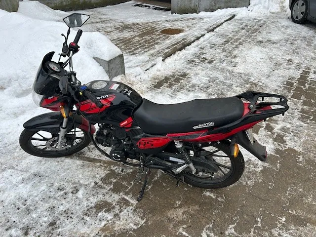 Czarno-czerwony motocykl marki Barton z napisem RS na boku, zaparkowany na śnieżnej nawierzchni. W tle widoczne są częściowo ośnieżone brukowane tory oraz fragment samochodu. Na siedzeniu widnieje napis Barton.