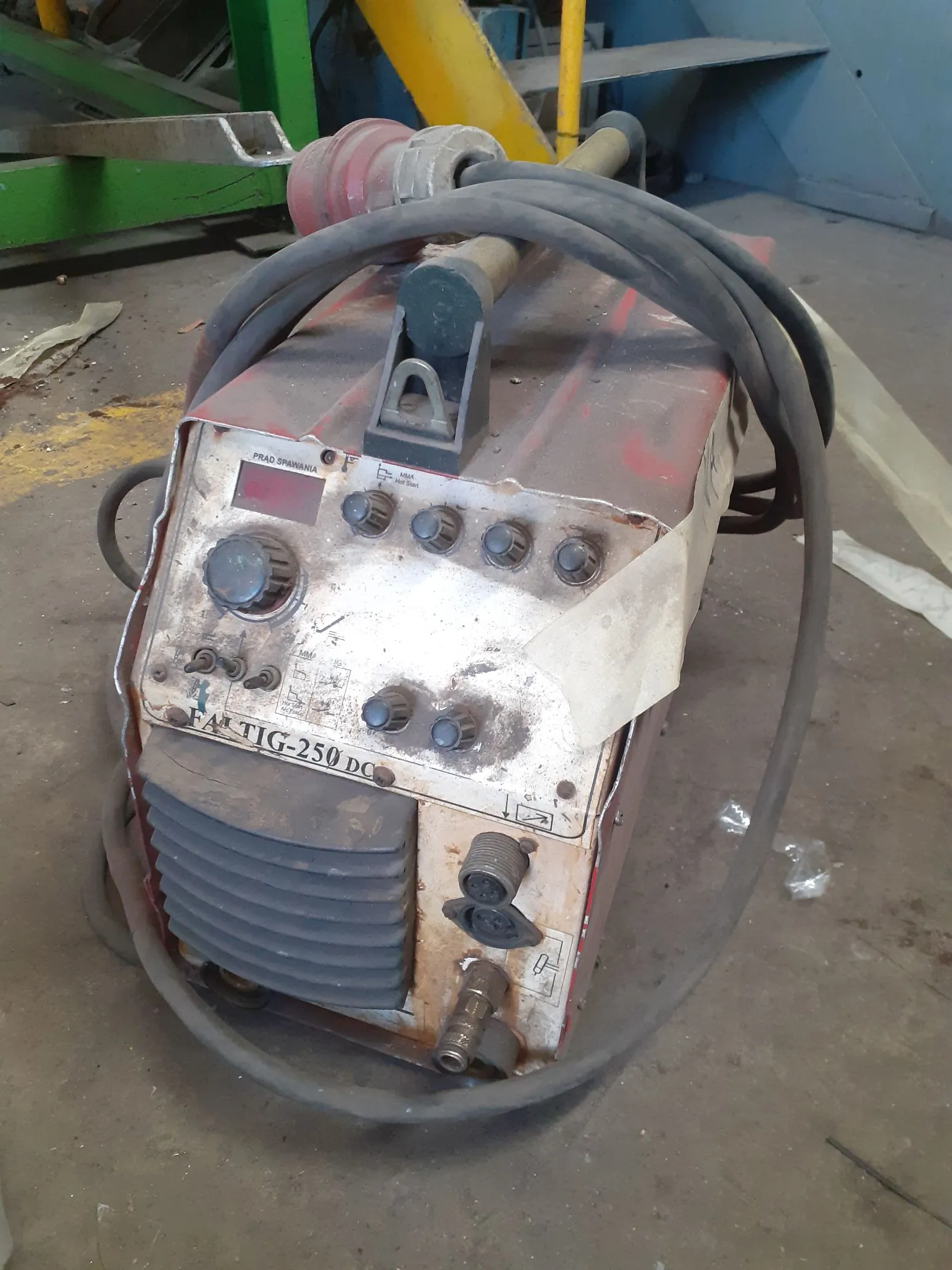 Stara, zabrudzona spawarka elektryczna w kolorze czerwonym i białym. Na przednim panelu widnieje napis 'TIG-250 DC'. Urządzenie posiada pokrętło regulacji, kilka przełączników oraz gniazda przyłączeniowe. Do maszyny podłączone są grube, czarne kable spawalnicze, które owijają się wokół obudowy.