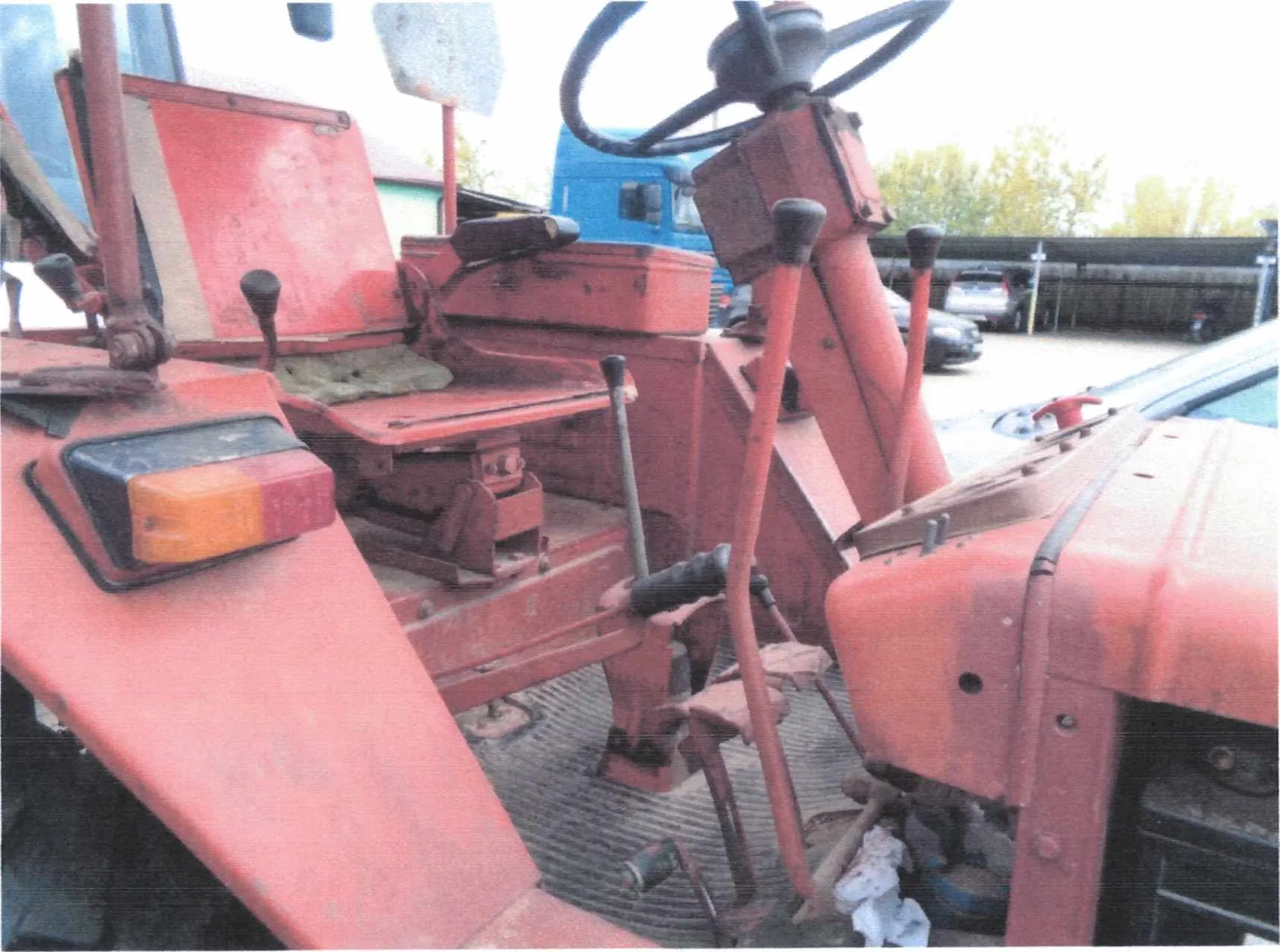 Zdjęcie przedstawia wnętrze czerwonego traktora. Widoczne są kierownica, siedzenie, dźwignie sterujące, reflektor z pomarańczowym i czerwonym segmentem oraz metalowa podłoga z siatką. W tle znajdują się niebieski ciężarówka, samochody osobowe, drzewa i budynki. Traktor wydaje się zużyty, z widocznymi śladami brudu i zużycia.