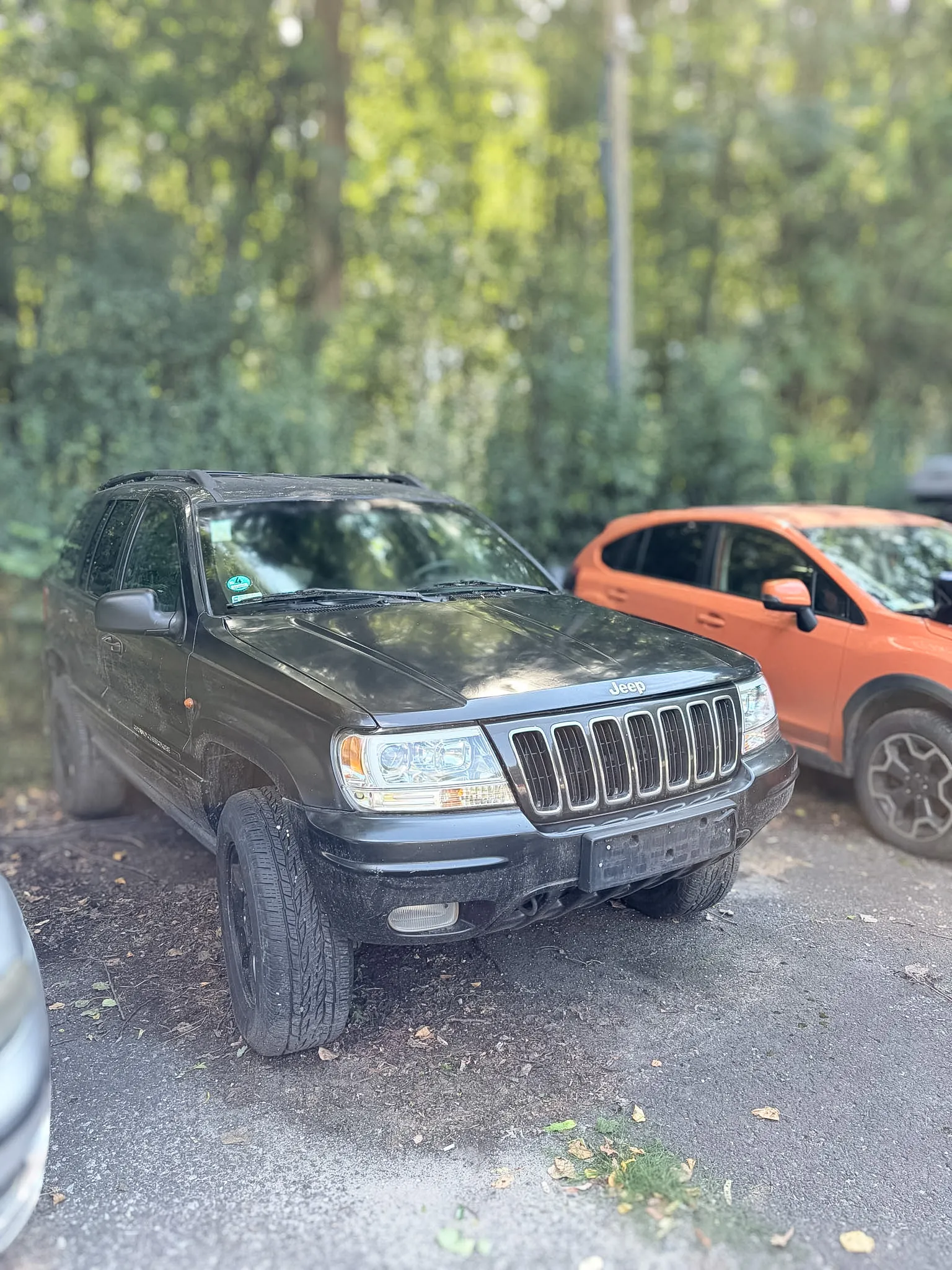 Czarny samochód terenowy marki Jeep zaparkowany na szutrowym poboczu obok pomarańczowego samochodu. W tle widoczne są zielone drzewa i krzewy.