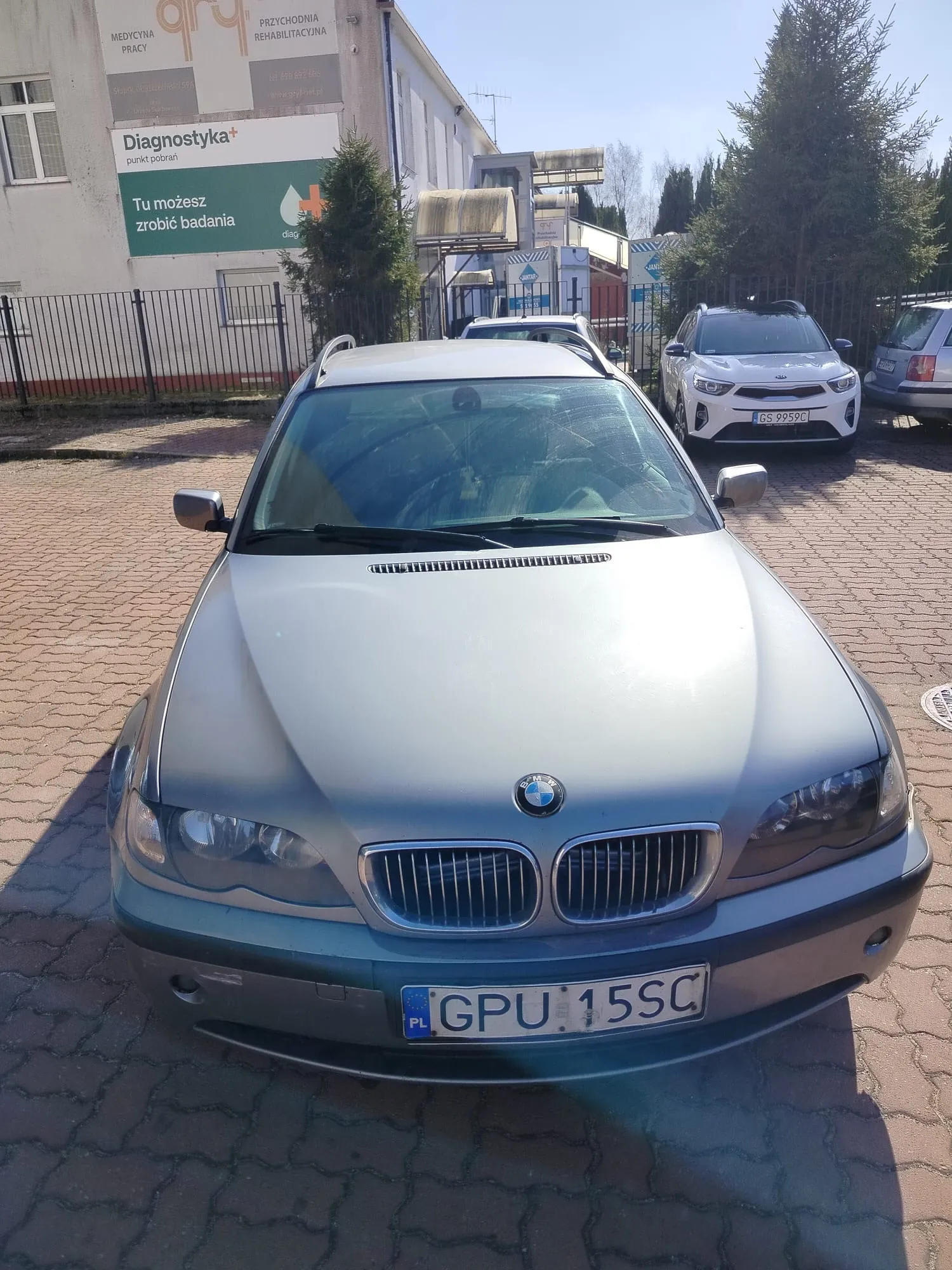 Przedni widok srebrnego samochodu marki BMW z tablicą rejestracyjną GPU 15SC (PL) zaparkowanego na brukowanej powierzchni. W tle budynek z napisami: 'Diagnostyka+ punkt pobrań', 'Tu możesz zrobić badania', 'MEDYCYNA PRACY', 'PRZYCHODNIA REHABILITACYJNA' oraz zielonym krzyżem. Widoczne są również inne pojazdy, w tym biały samochód Kia z tablicą GS 9959C, ogrodzenie metalowe i drzewa. Niebo jest jasne, pogoda słoneczna.