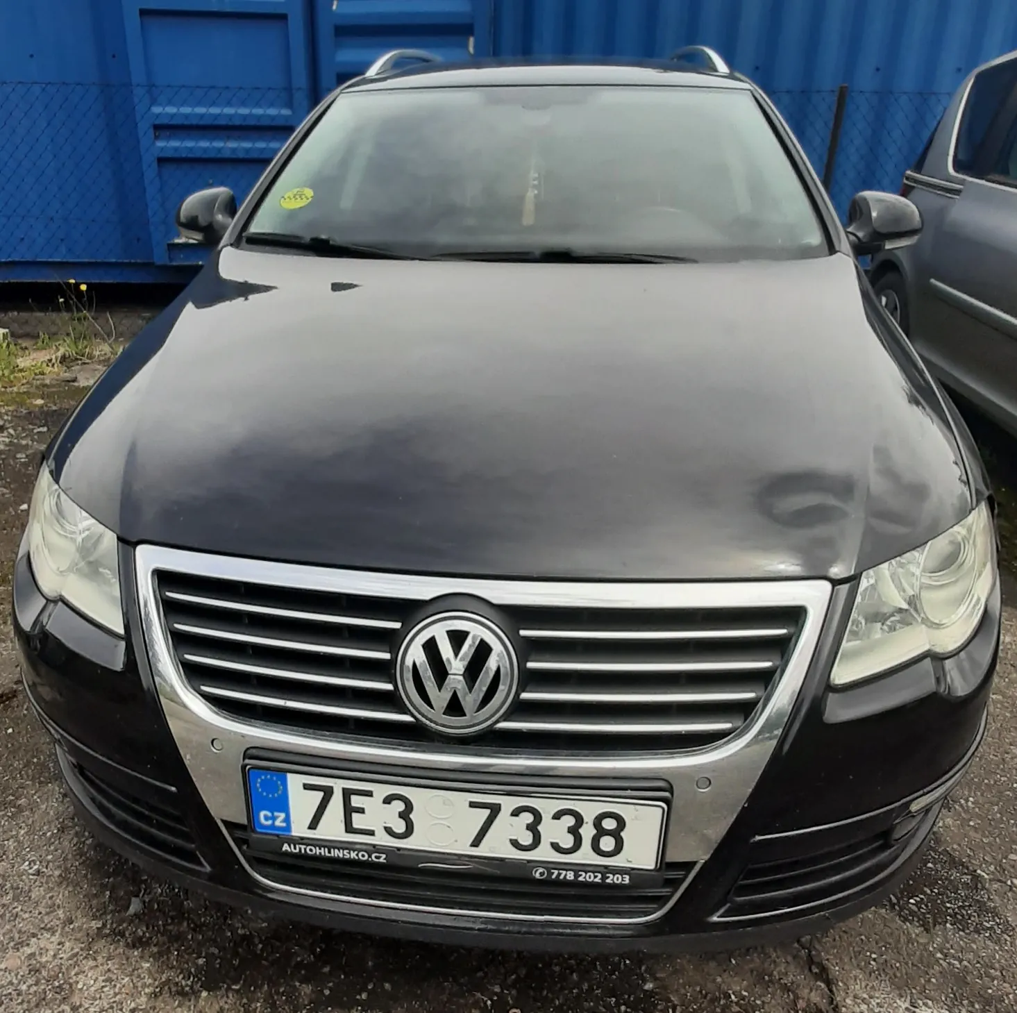 Czarny samochód marki Volkswagen z przodu, z widocznym logo na masce. Tablica rejestracyjna z napisem 7E3 7338, przedrostek CZ oraz napisem AUTOHLINSKO.CZ i numerem telefonu 778 202 203 pod tablicą. Tło stanowi niebieski kontener i siatka ogrodzeniowa. Po prawej stronie częściowo widoczny inny samochód. Samochód stoi na podłożu z drobnej kostki.