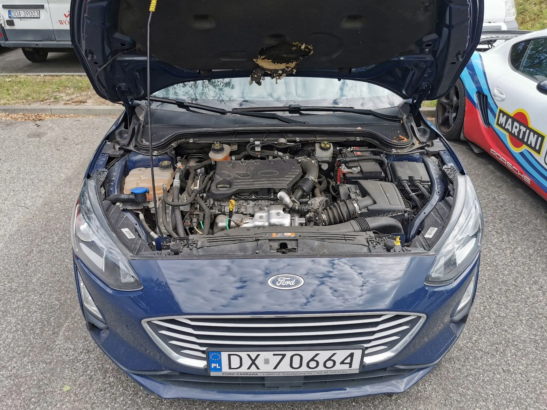 Widok z przodu na ciemnoniebieski samochód marki Ford z otwartą maską, ukazującą silnik i podzespoły mechaniczne. Na przednim zderzaku znajduje się tablica rejestracyjna o numerze DX 70664. W tle po prawej stronie widoczny jest fragment białego samochodu z logo Martini i napisem Porsche.